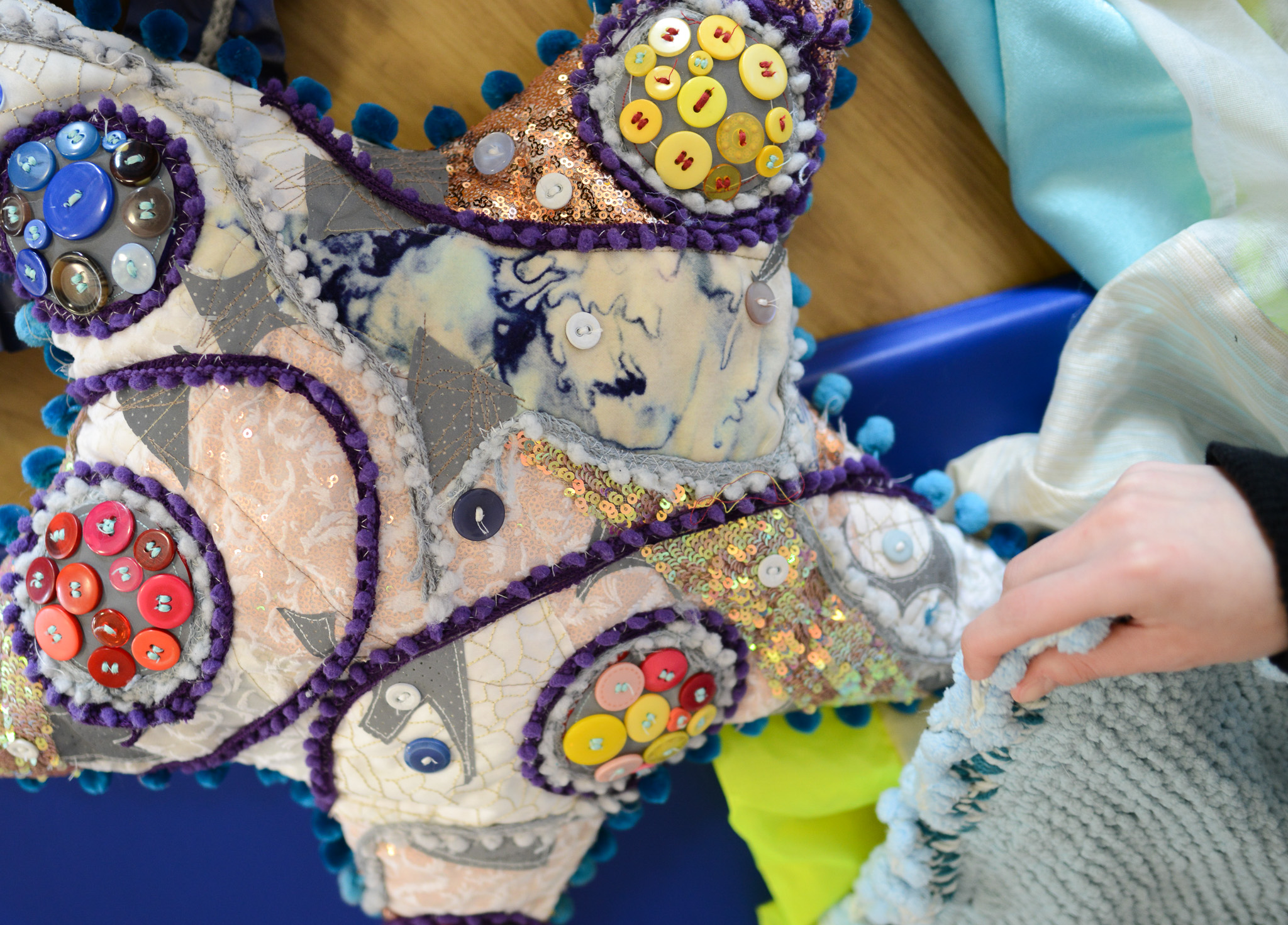 Colourful, textured star-shaped pillow with buttons and sequins, held by a hand, on a blue and wooden background.