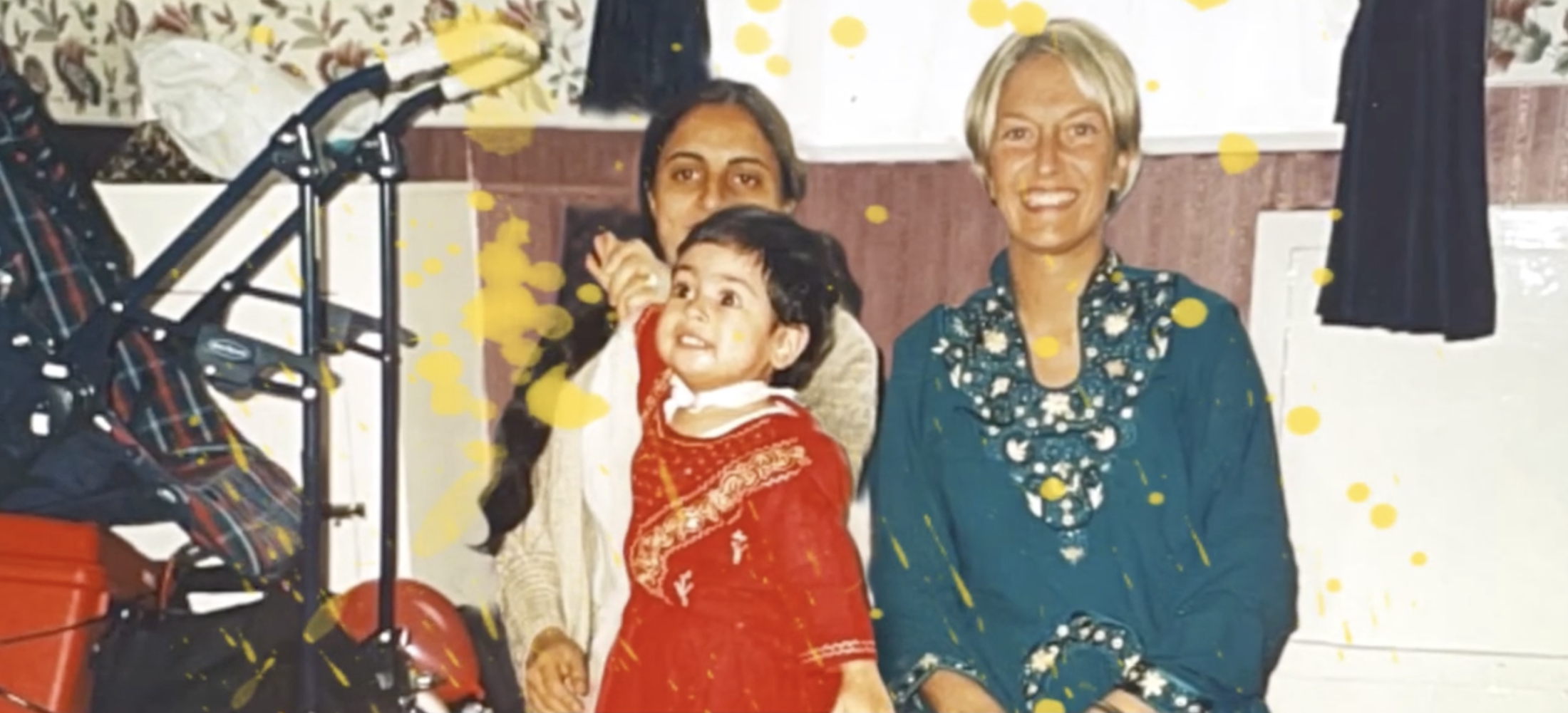 A young child in a red traditional outfit stands between two women, one in a dark dress and the other in a teal dress, all smiling indoors with a patterned wall and yellow spots overlaying the photo.