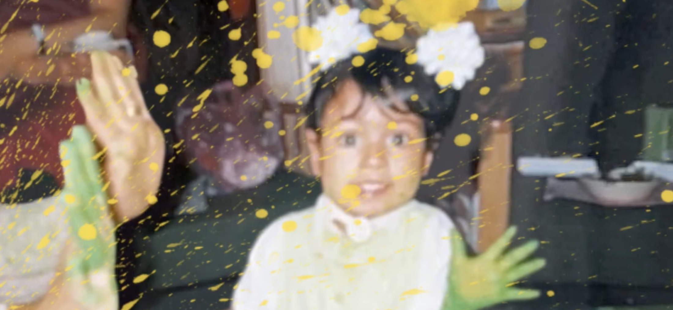 A young child smiles excitedly, with green hands raised and splattered with yellow paint. Bright flowers adorn their hair.