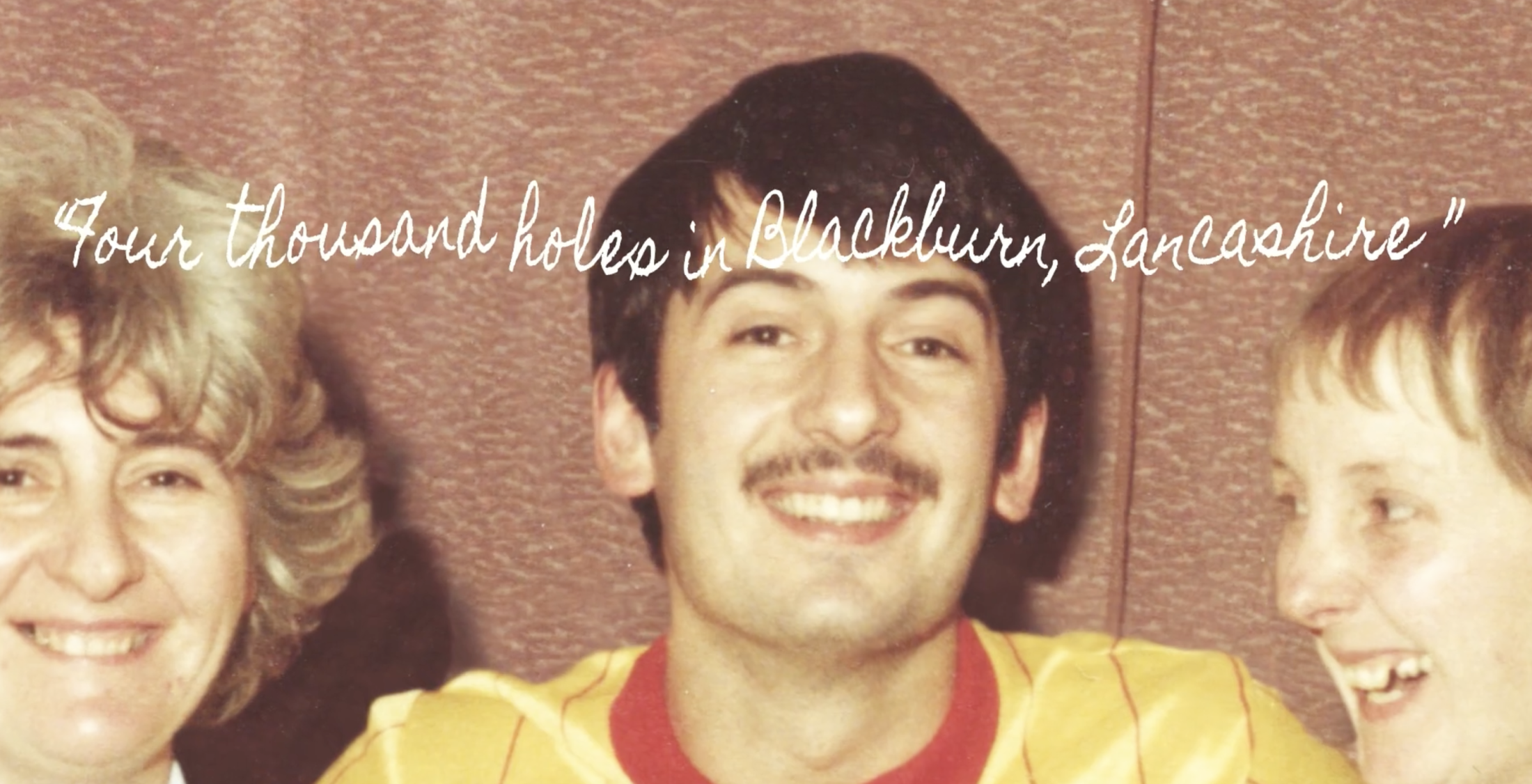 A smiling man with a moustache in a yellow and red shirt is surrounded by two joyful women. Text reads 