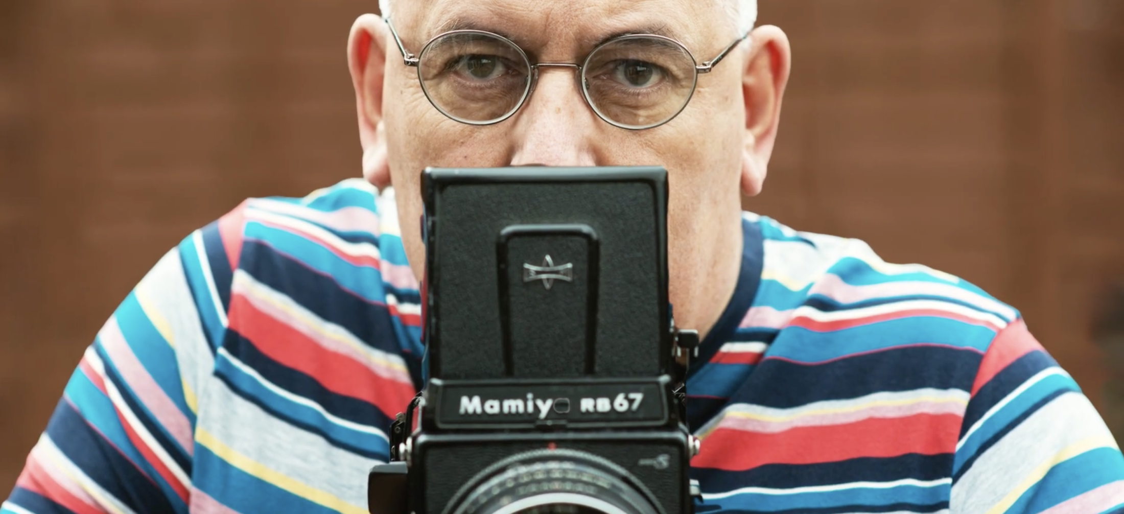 A person wearing glasses and a colourful striped shirt is holding a Mamiya RB67 camera. The background is softly blurred.