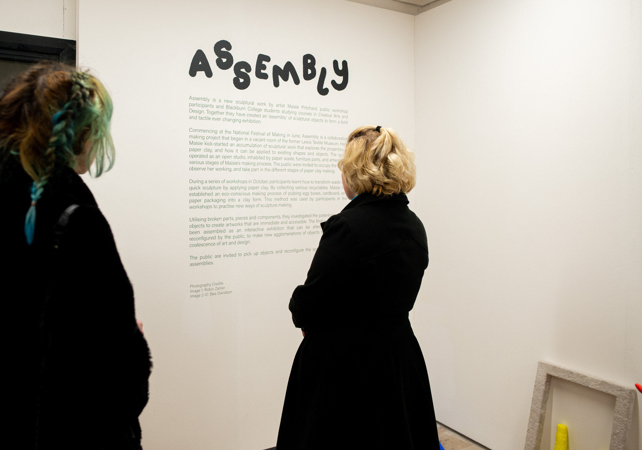 Two people stand in a gallery, reading a wall text titled 