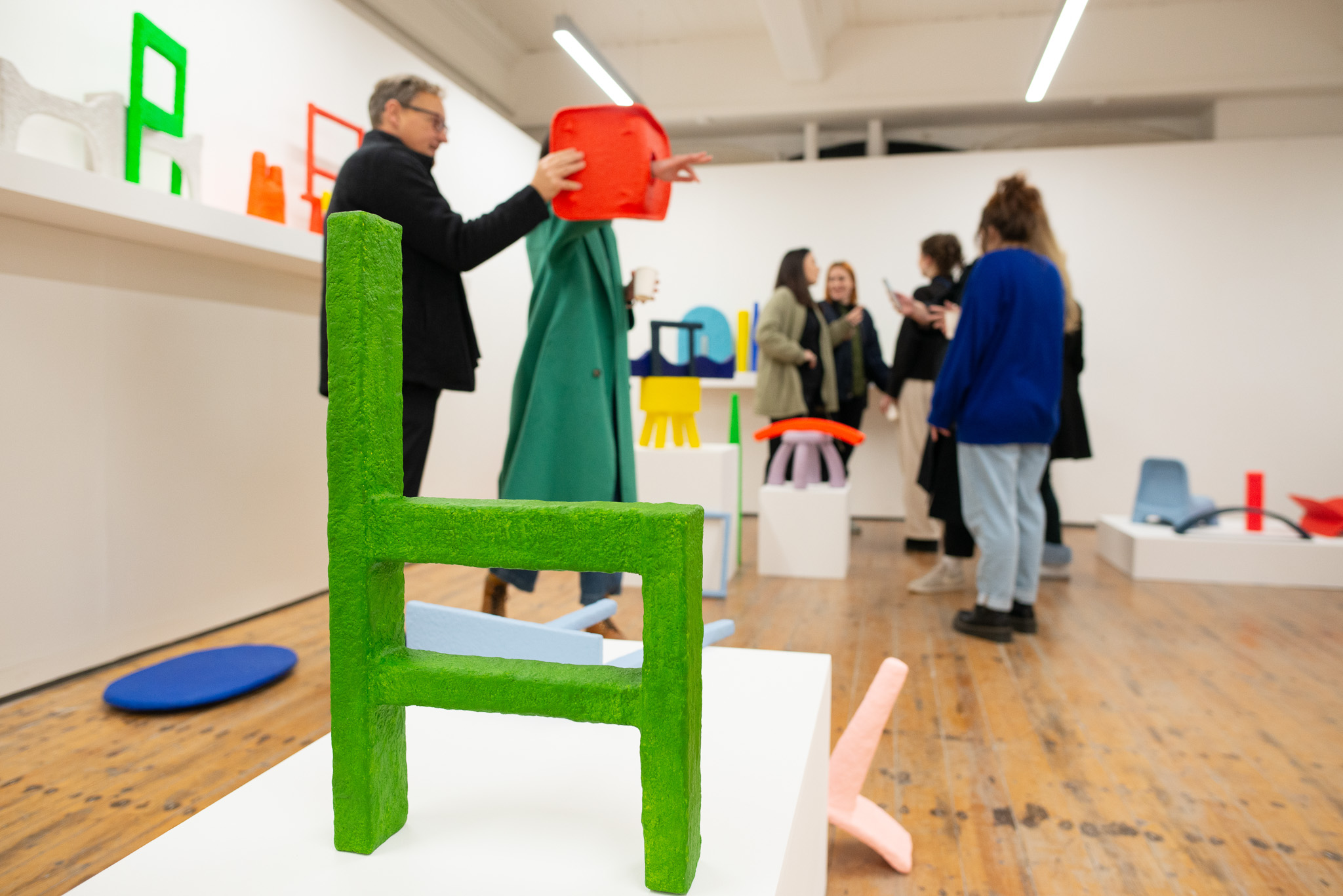 A group of people talk in a brightly lit art gallery featuring colourful, abstract chairs on white plinths; a vivid green chair is in the foreground.