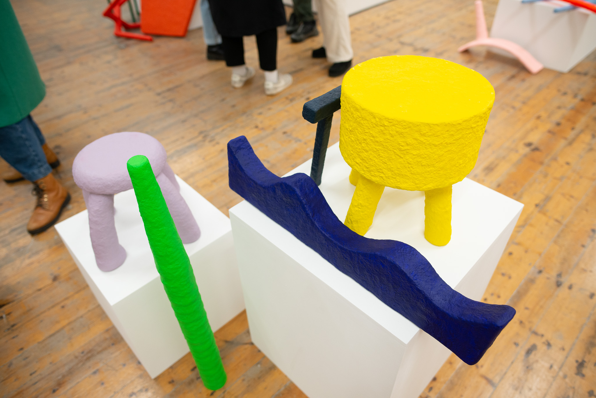 Colourful abstract sculptures on white pedestals in a gallery; a yellow table with a blue wavy bar and a purple stool with a green pole on a wooden floor.