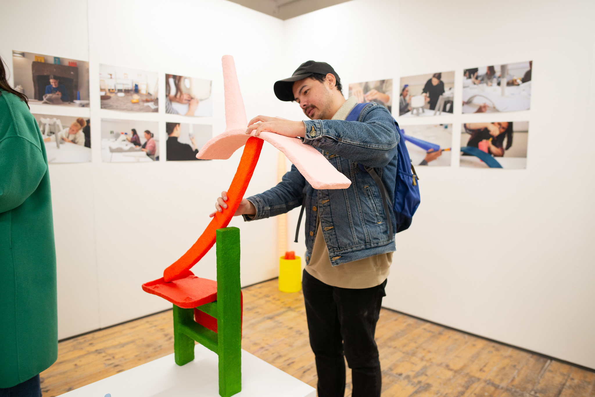 A person in a denim jacket and black cap interacts with a colourful abstract sculpture in a gallery, with photo prints on the walls behind.
