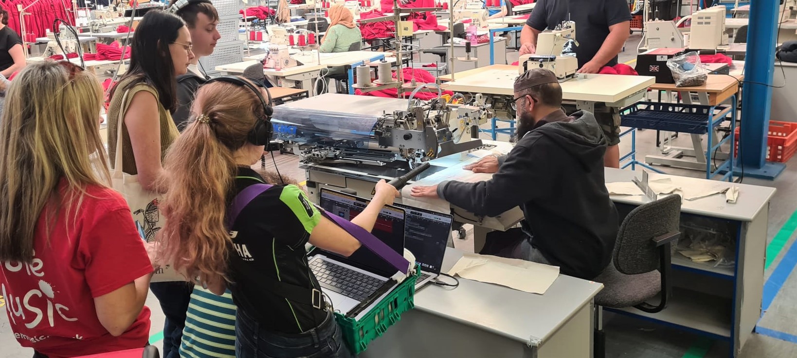 Several people stand around a seated man operating an industrial sewing machine in a busy workshop, observing and possibly learning the process. Desks, fabric, and sewing equipment are visible throughout the space.