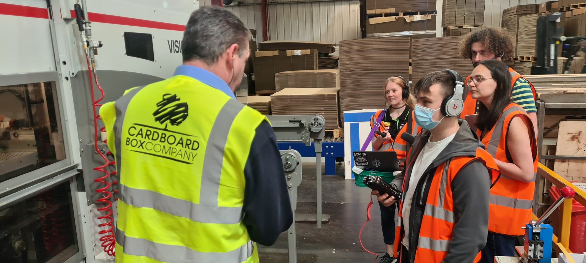 A man in a yellow Cardboard Box Company vest gives a tour to four young people wearing orange vests and headsets in a factory filled with cardboard and machinery.