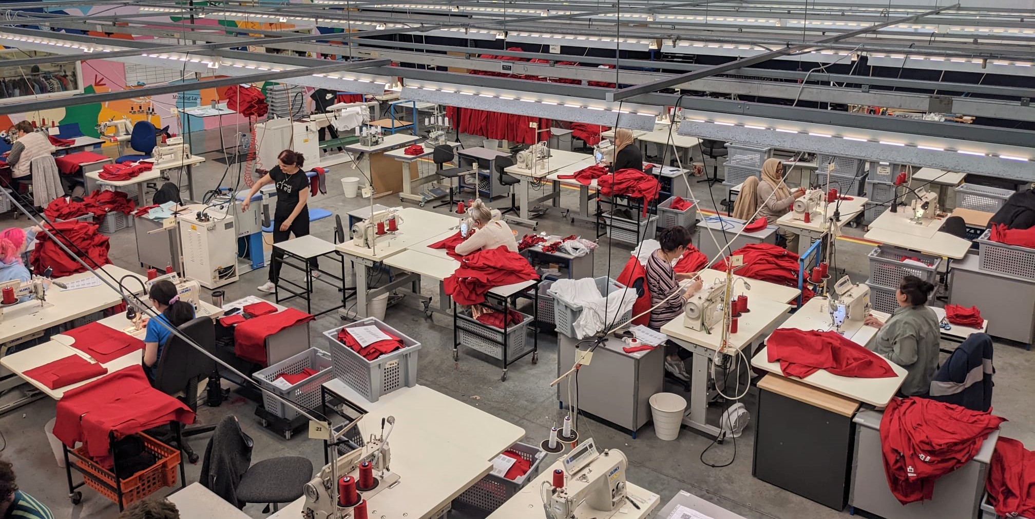A view of a garment factory with workers sitting at sewing machines, stitching bright red fabric. The workspace is organised in rows, with tables, equipment, and overhead lights.
