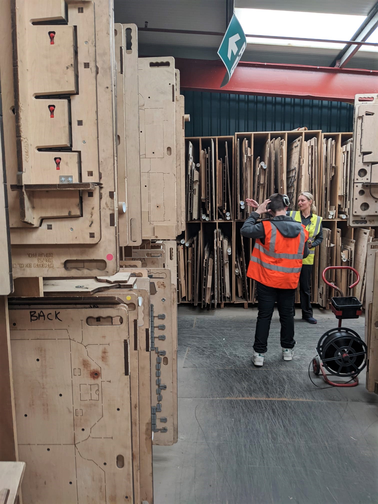Two people in high-visibility vests stand in a warehouse filled with large wooden templates or moulds stored vertically on racks. One person gestures while the other listens. A green exit sign hangs above.