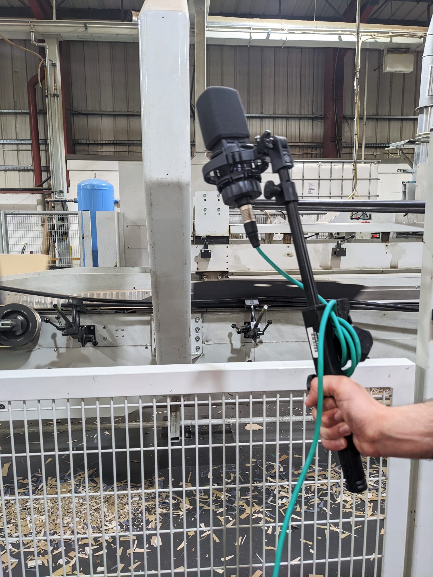 A person holds a microphone on a stand with a green cable in front of industrial machinery inside a factory setting, with metal fencing and wood shavings on the floor.