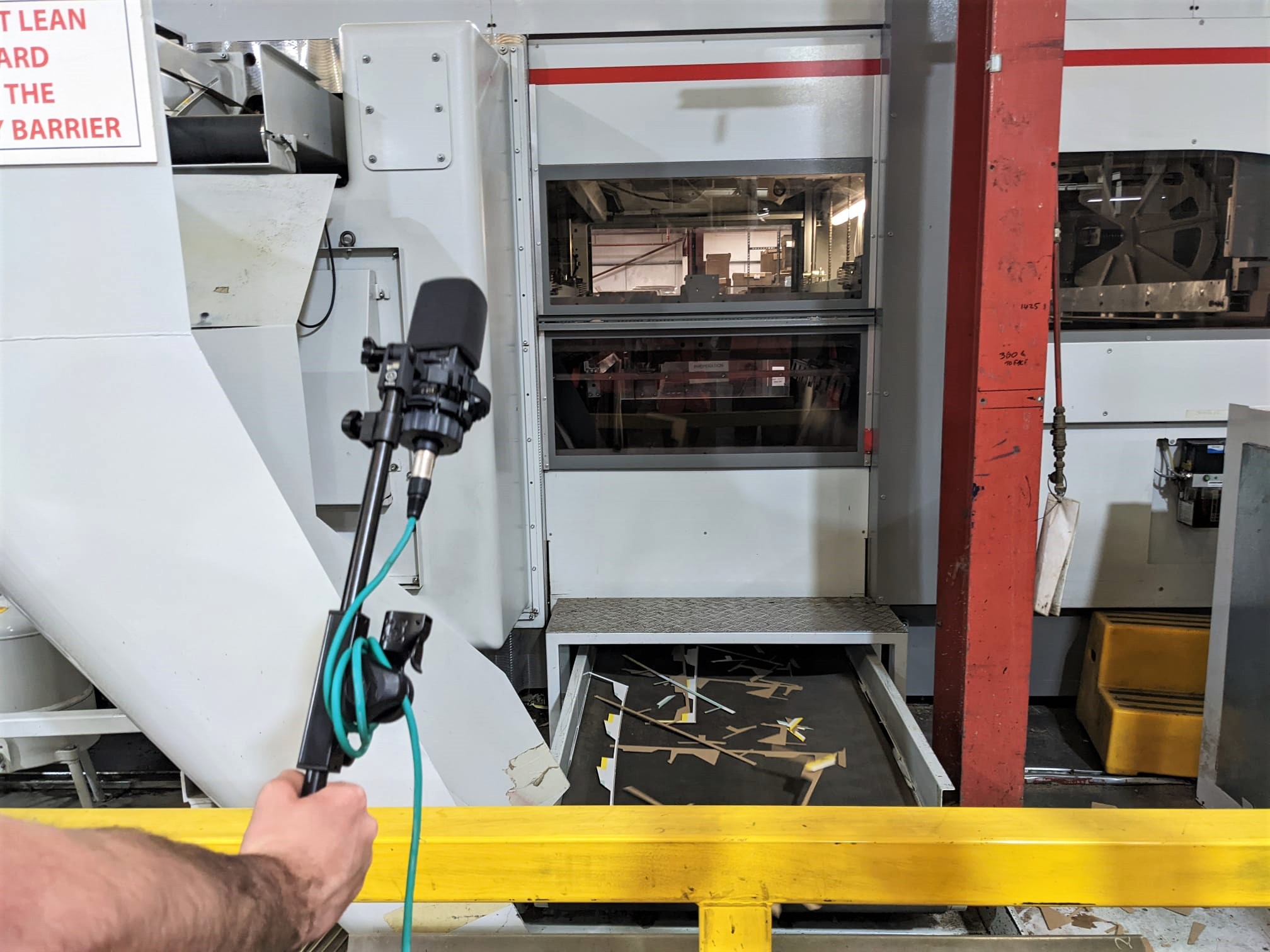 A person holds a microphone on a stand near a large industrial machine with scattered wooden cut-outs on the floor, behind a yellow safety barrier.
