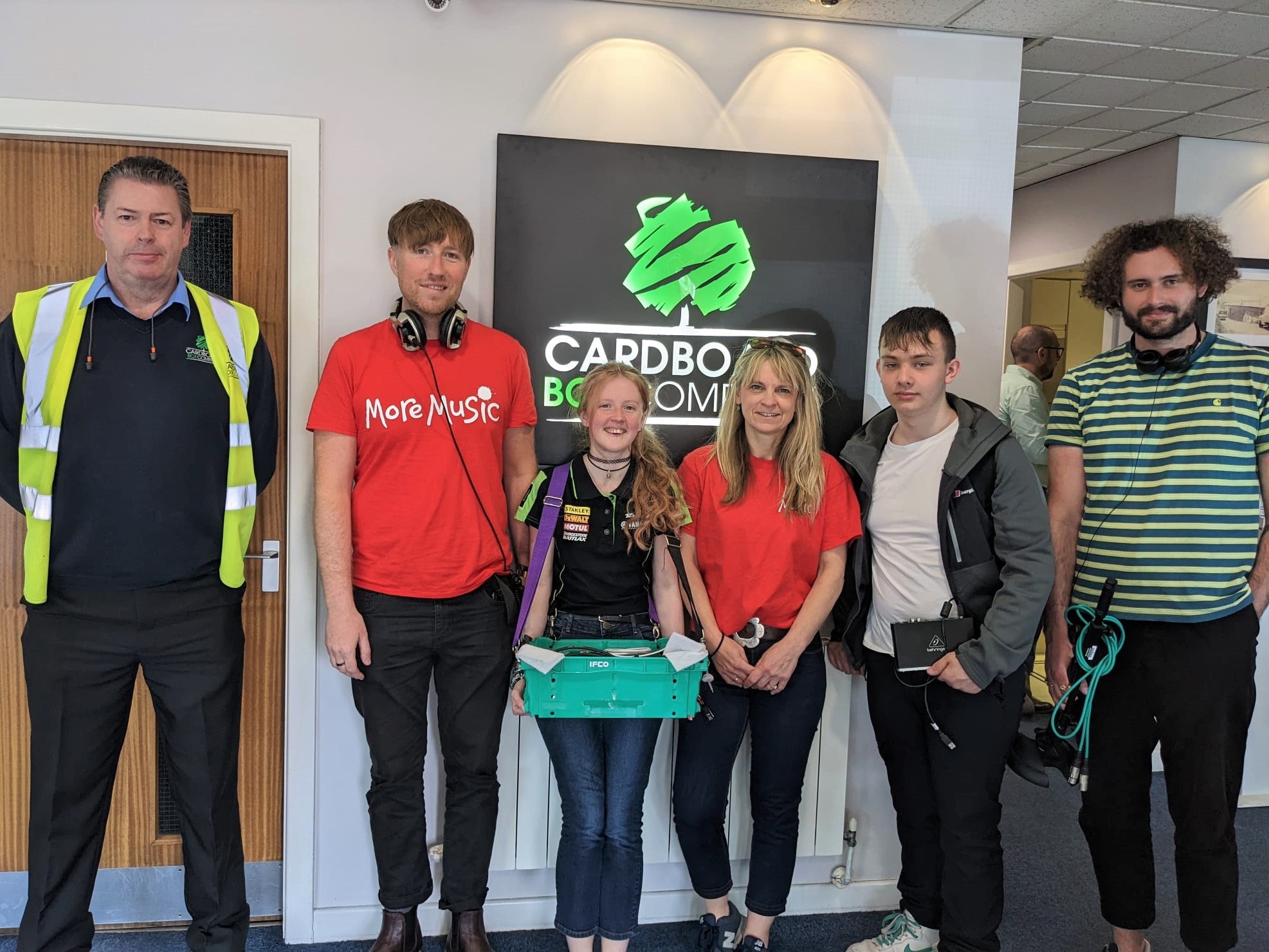 Six people stand indoors in front of a sign that reads CARDBOARD BOX COMPANY. One person wears a high-visibility vest, two wear red shirts, and the group is smiling or looking at the camera.