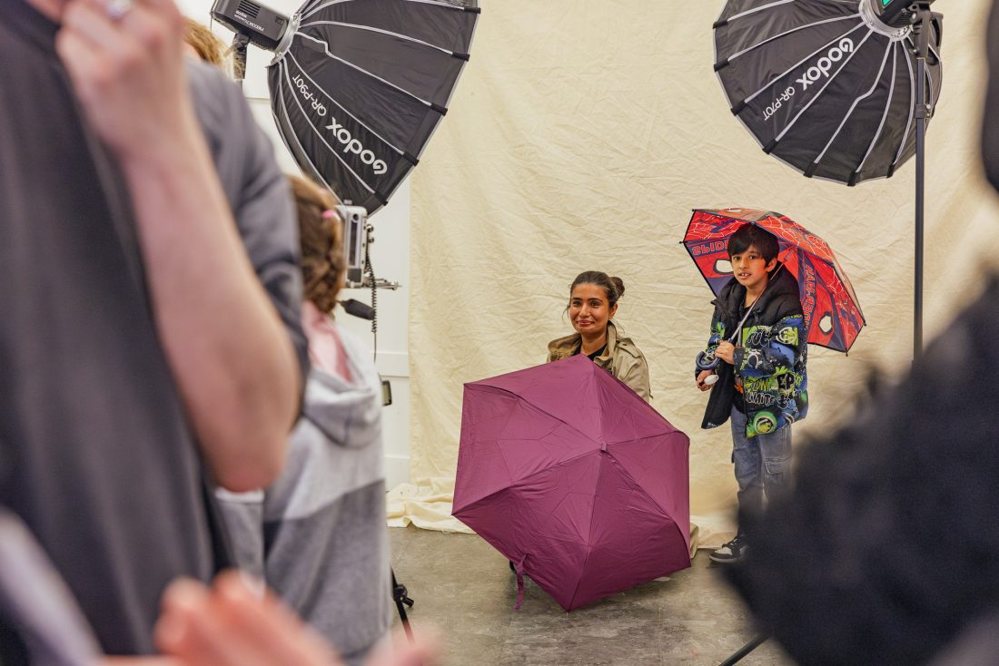 A woman sits behind a purple umbrella, smiling, whilst a child beside her holds a colourful umbrella, both under studio lights with people visible in the foreground. The backdrop is a plain beige fabric.