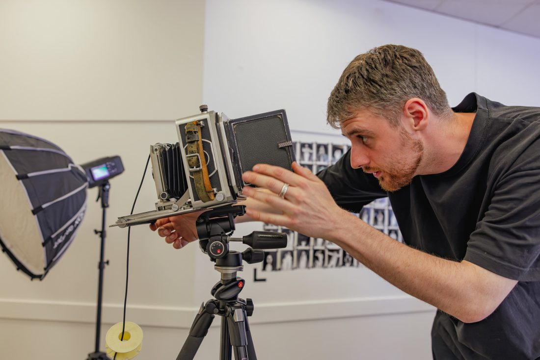 A man adjusts a vintage large format camera on a tripod in a photography studio, with lighting equipment and black-and-white portraits visible in the background.