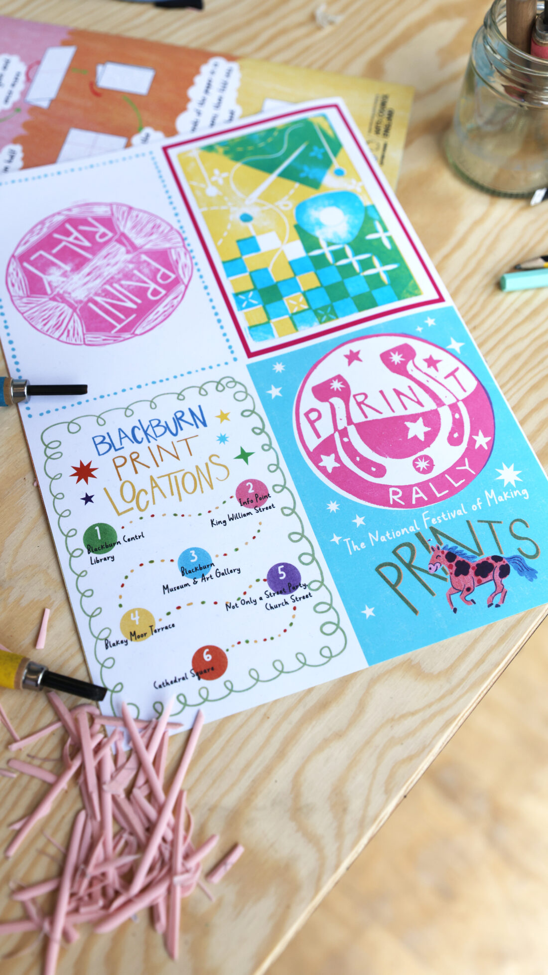 Three colourful festival flyers on a wooden table, surrounded by pink pencil shavings and drawing tools. The flyers feature event names, a map, and playful illustrations in blue, pink, green, and yellow.