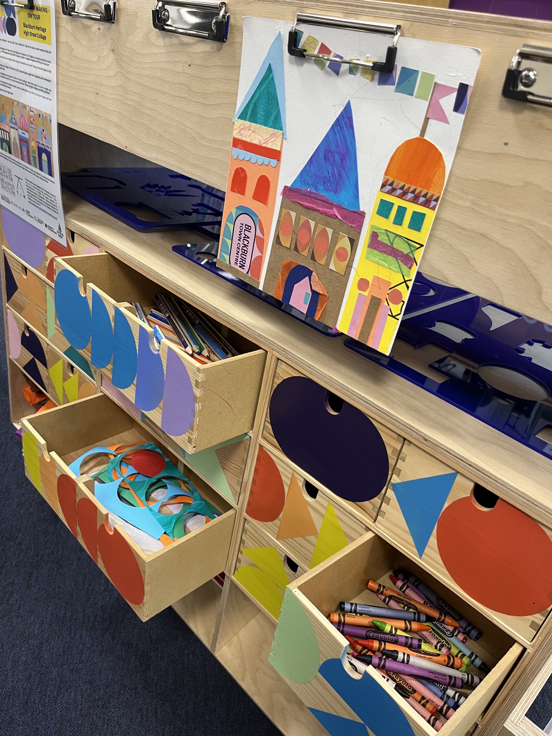 A wooden art station with open drawers filled with colourful paper shapes and crayons. On top, a clipboard displays a vibrant drawing of geometric buildings in various colours and patterns.
