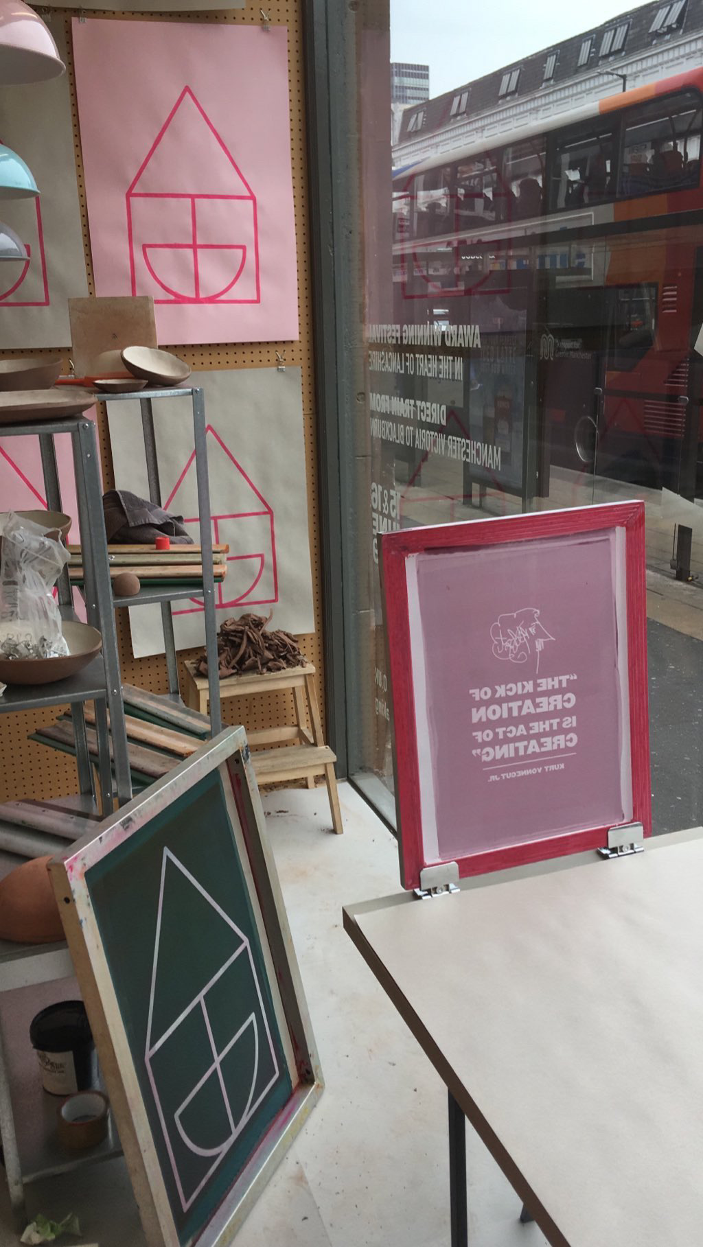 A pottery studio with shelves of ceramics and tools beside a window. Colourful prints featuring simple house shapes are displayed. Outside, a red double-decker bus passes by on a city street.