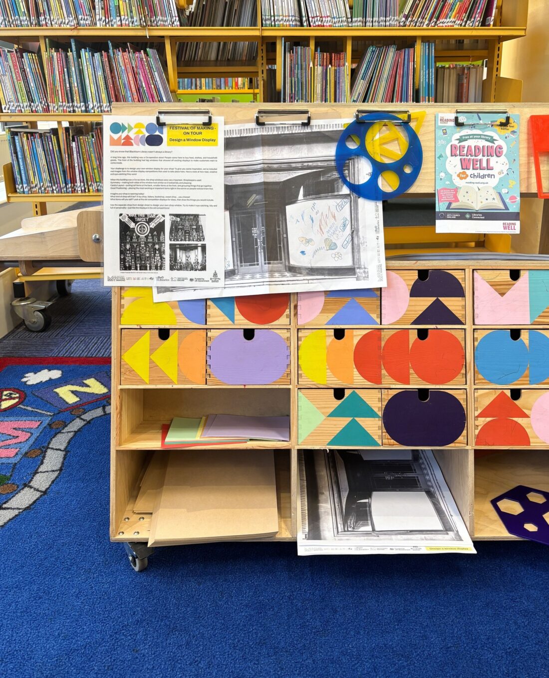 A colourful classroom with bookshelves, art supplies, and storage drawers decorated with geometric patterns. Large printed posters, a blue and yellow stencil, and a Reading Well flyer are visible on the wooden unit.