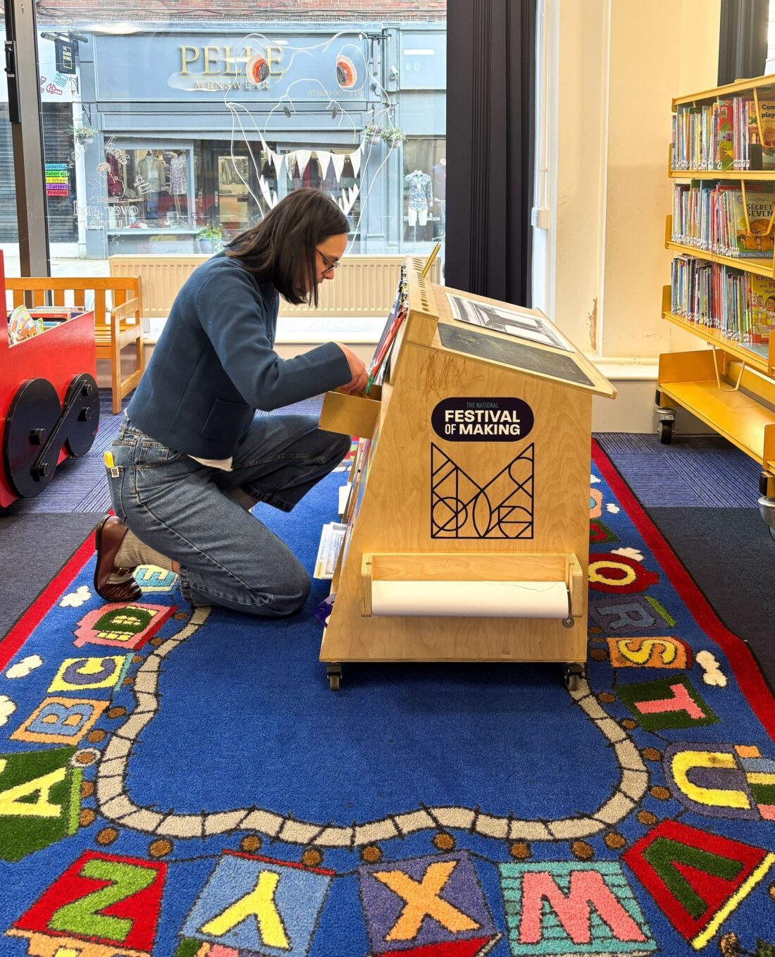 A person kneels on an alphabet rug in a library, organising books on a wooden display labelled Festival of Making. Shelves of colourful books and a toy trolley are visible in the background.