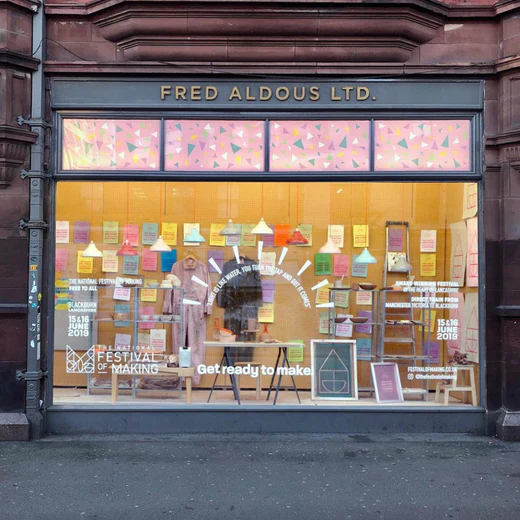 Shopfront of Fred Aldous Ltd. with a colourful display of posters, crafts, and supplies in the window. The text Get ready to make and festival dates are visible among the creative decor.
