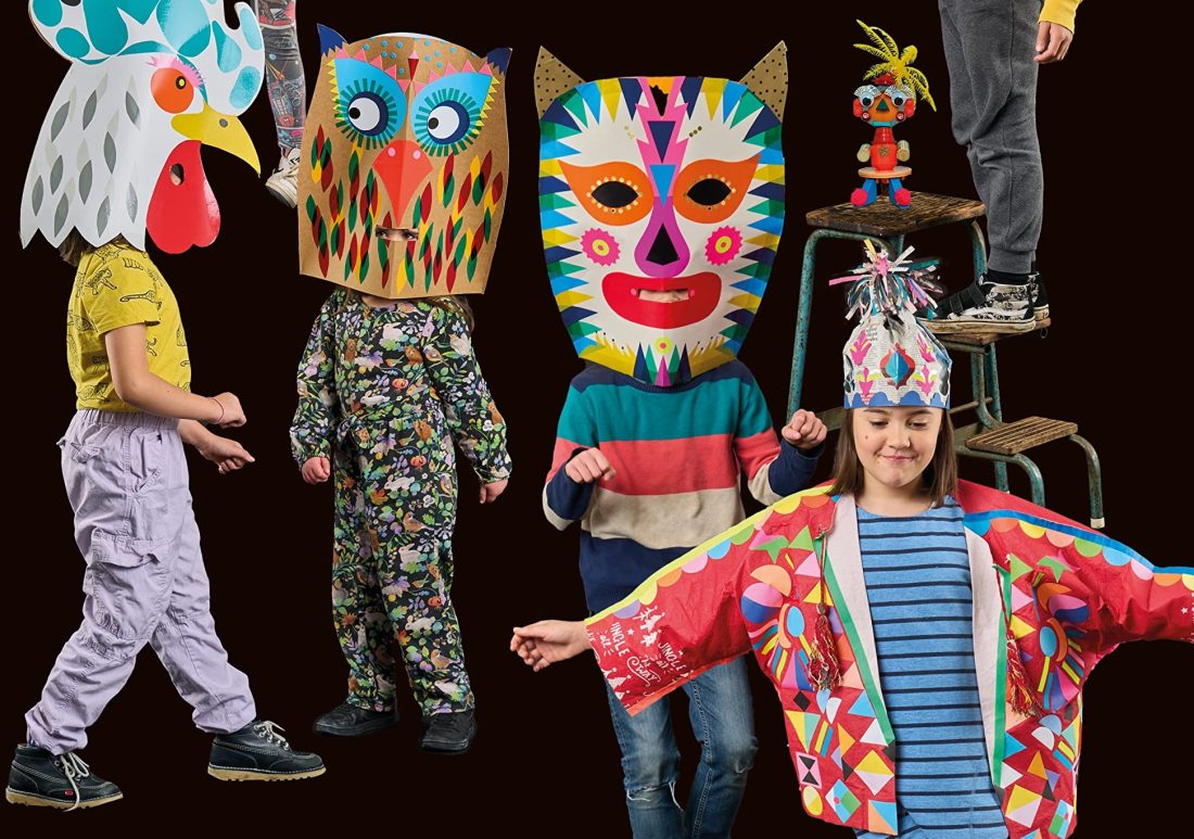 Children wearing colourful, animal-inspired paper masks, hats, and costumes stand and pose playfully against a dark background. One child stands on a chair, while another wears a vibrant jacket and decorated hat.