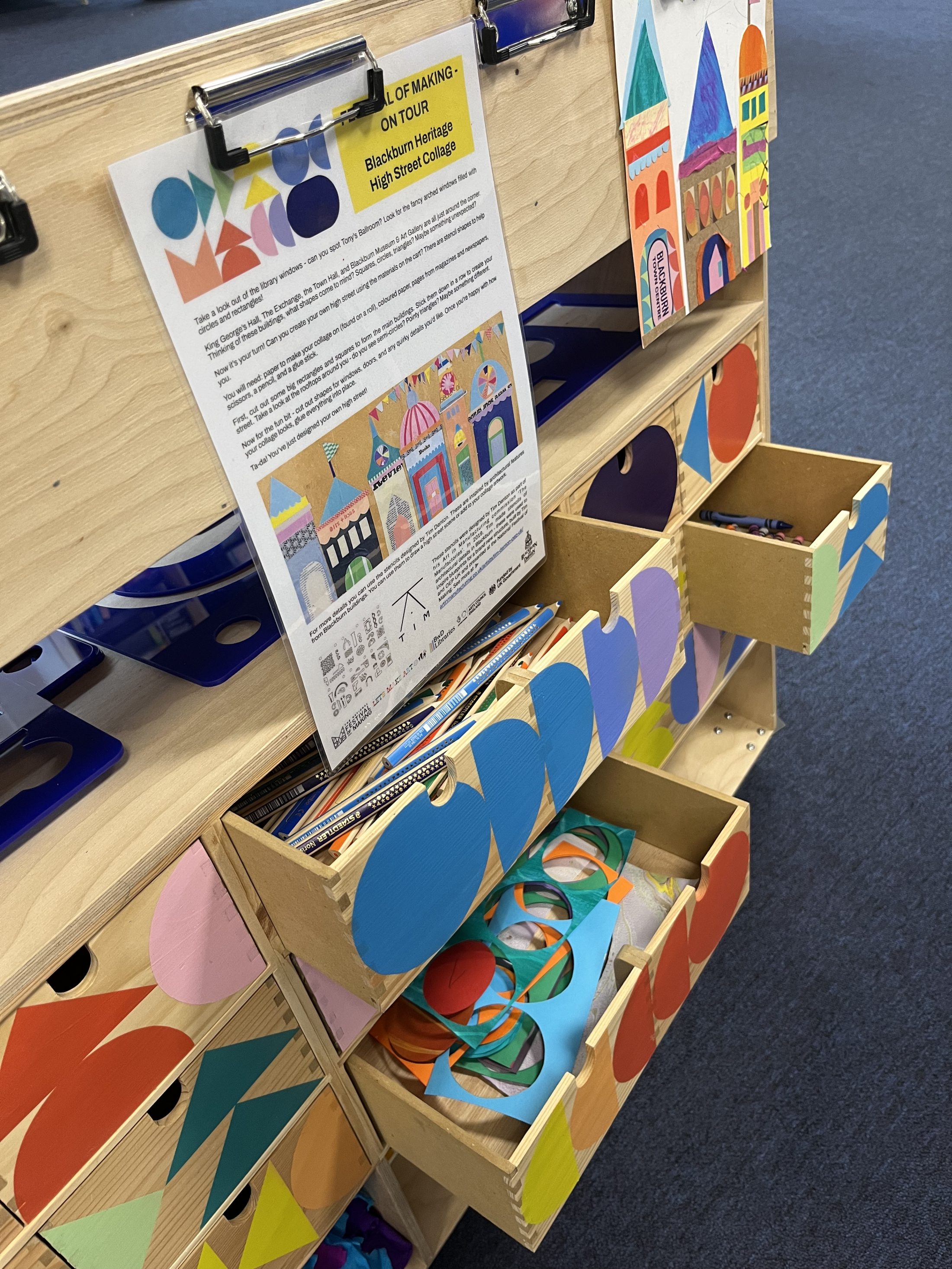A wooden organiser with open drawers holds colourful paper shapes and crafts. A printed instruction sheet is attached above, and vibrant, geometric decorations cover the front of the organiser. The setting appears to be a classroom or creative space.