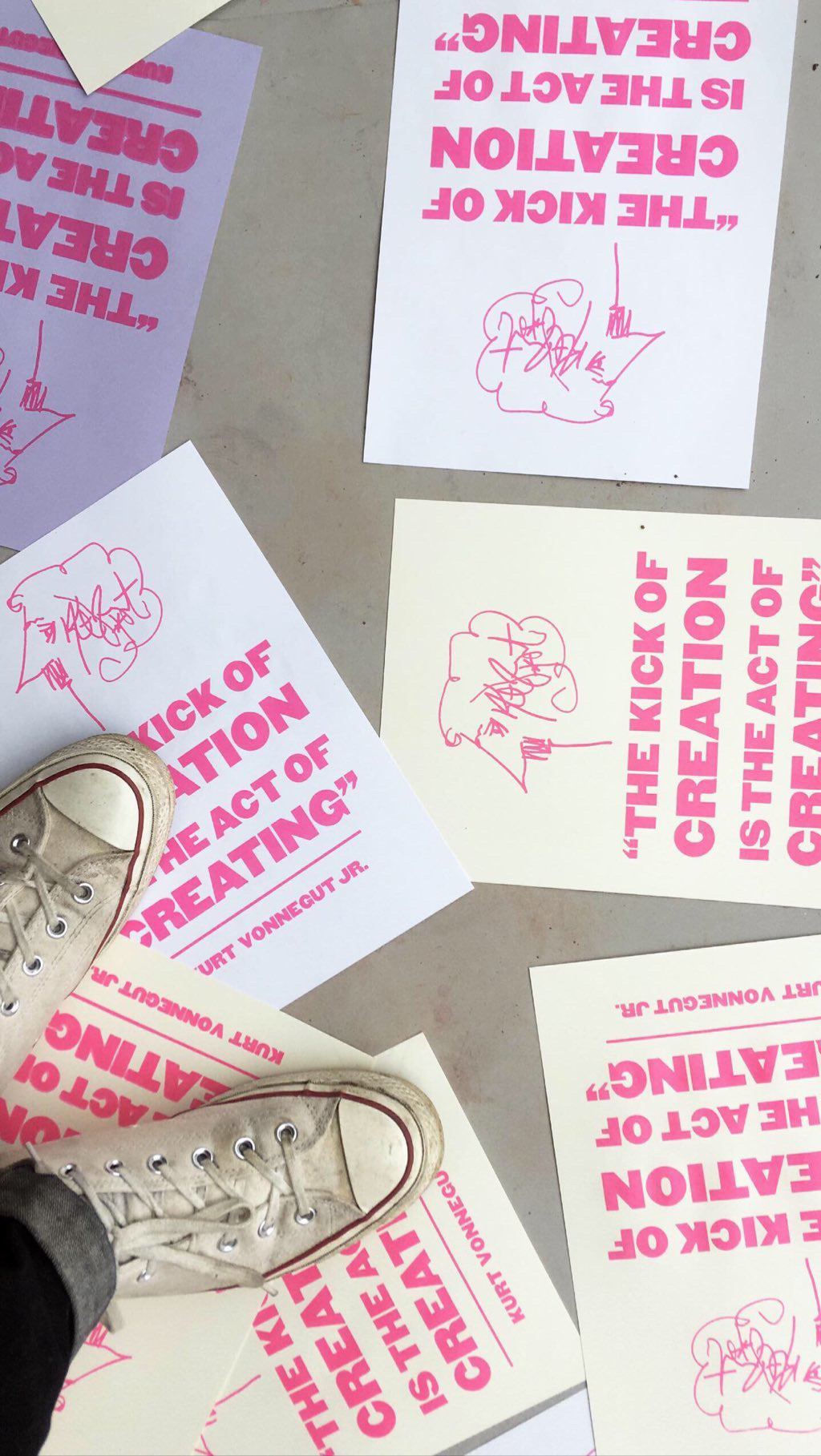 A person wearing worn white Converse trainers stands among scattered posters on the floor. The posters read “THE KICK OF CREATION IS THE ACT OF CREATING” in bold pink letters with abstract doodles.