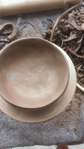 A person smooths the surface of a clay bowl with their fingers on a textured mat, surrounded by clay scraps and pottery tools.