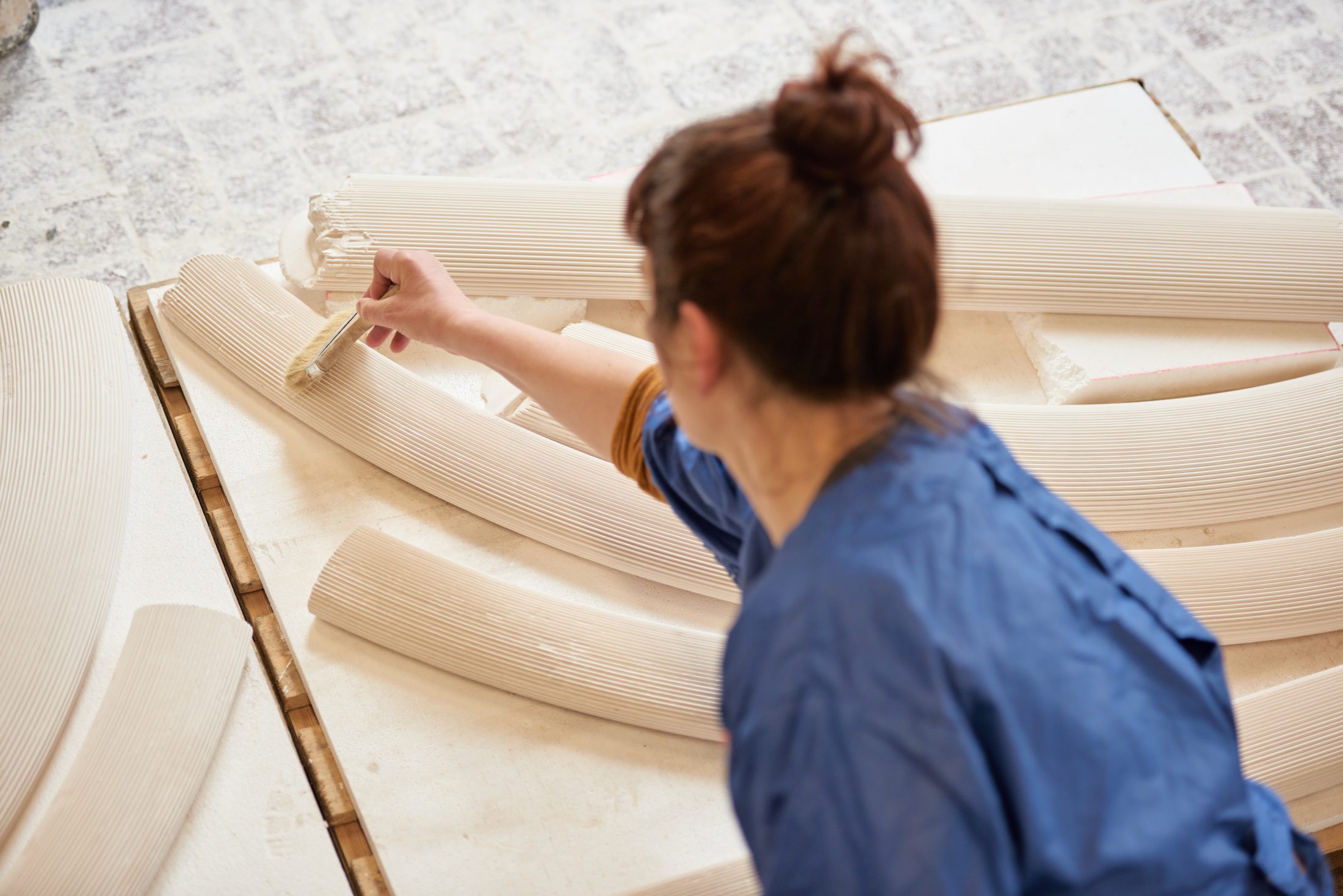 A person with brown hair in a bun, wearing a blue shirt, uses a brush to work on large, curved, ribbed ceramic pieces arranged on the floor. The surface below is textured and light coloured.