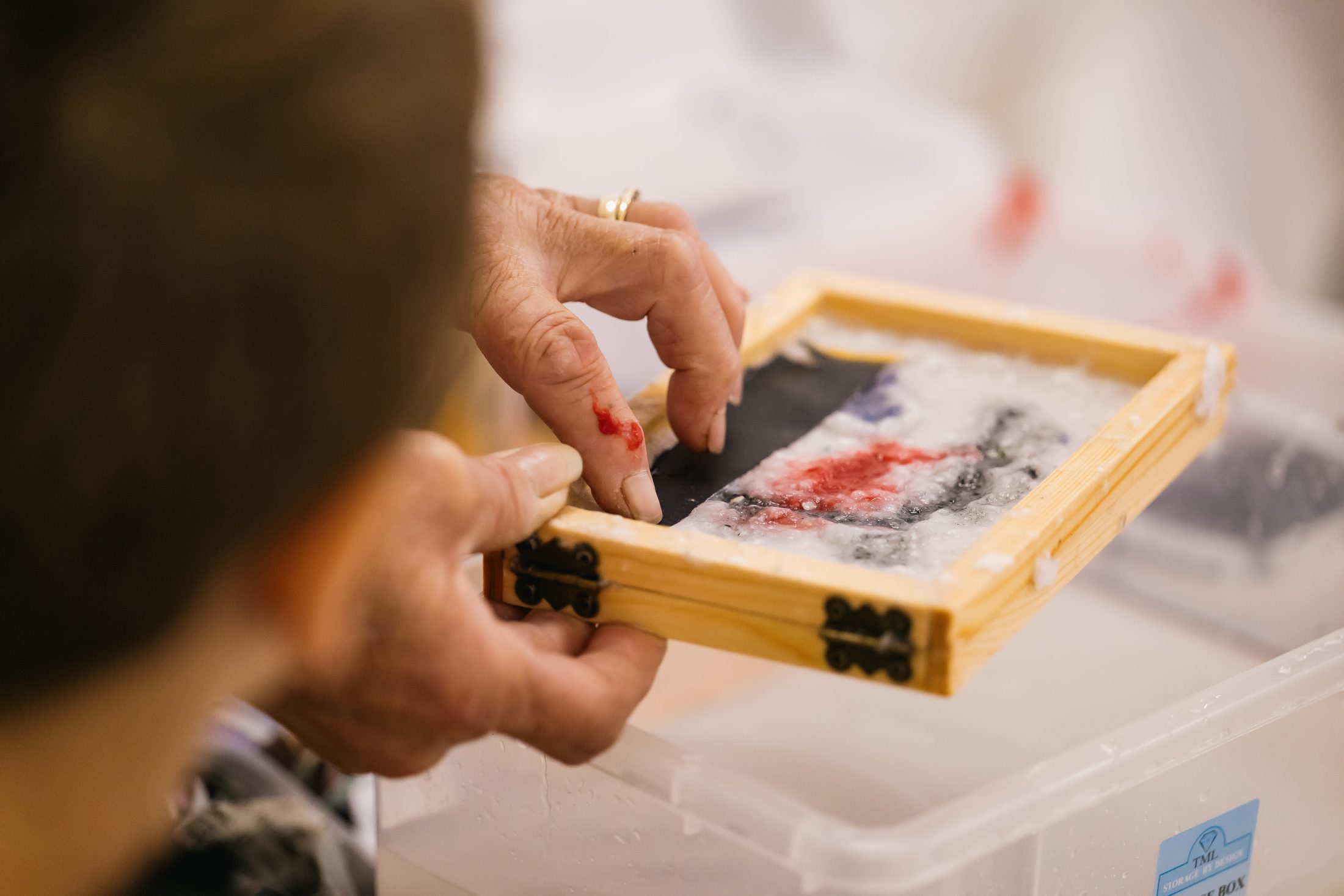 A person’s hands work on a small papermaking frame filled with wet pulp, red and black pigments, over a plastic bin. Another person observes closely in the foreground.
