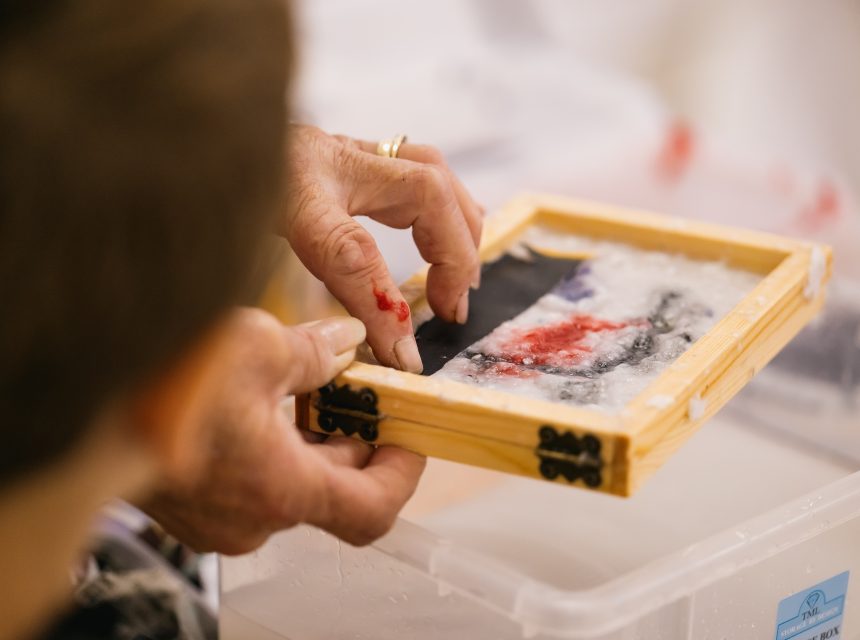 A person’s hands work on a small papermaking frame filled with wet pulp, red and black pigments, over a plastic bin. Another person observes closely in the foreground.
