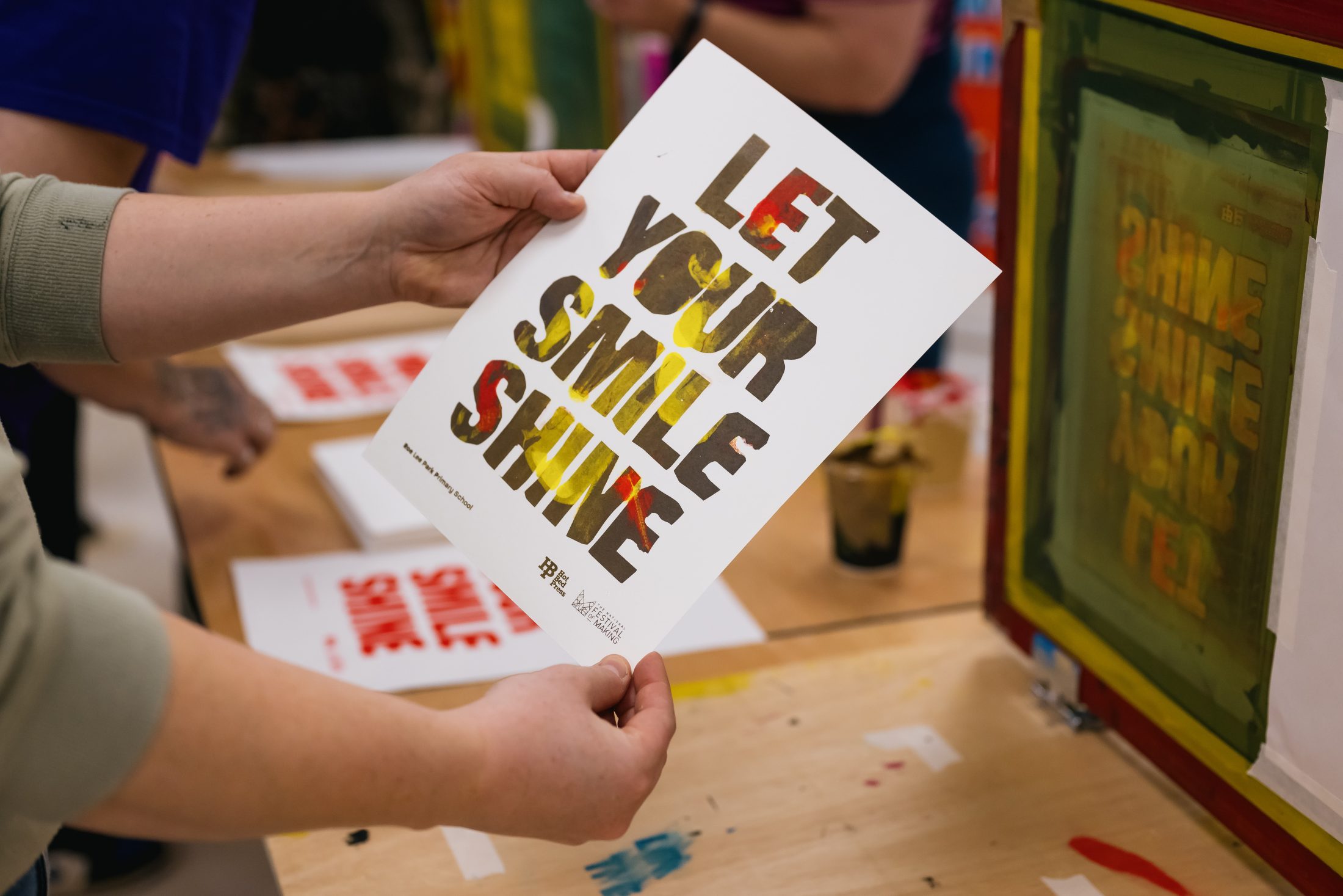 A person holds a printed poster that says, “LET YOUR SMILE SHINE” in bold, colourful letters, with screen printing materials and another similar print visible on the table.