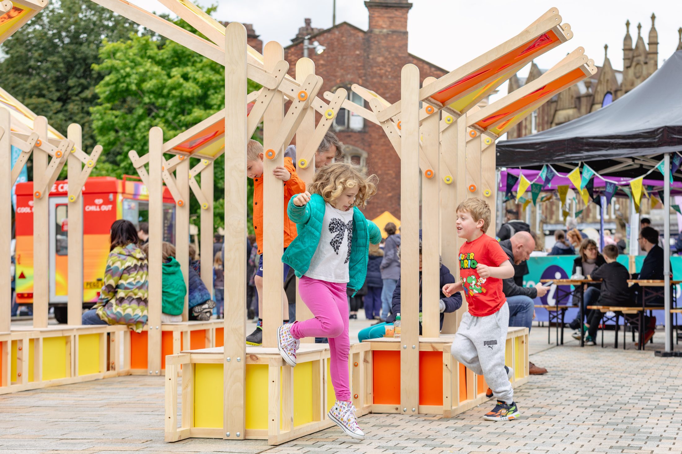 Two children play and smile among colourful wooden structures in an outdoor public space, whilst adults sit at tables and others enjoy the lively atmosphere in the background.