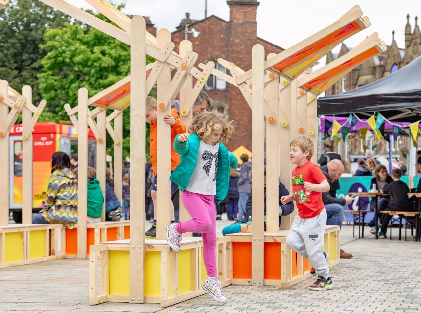 Two children play and smile among colourful wooden structures in an outdoor public space, whilst adults sit at tables and others enjoy the lively atmosphere in the background.