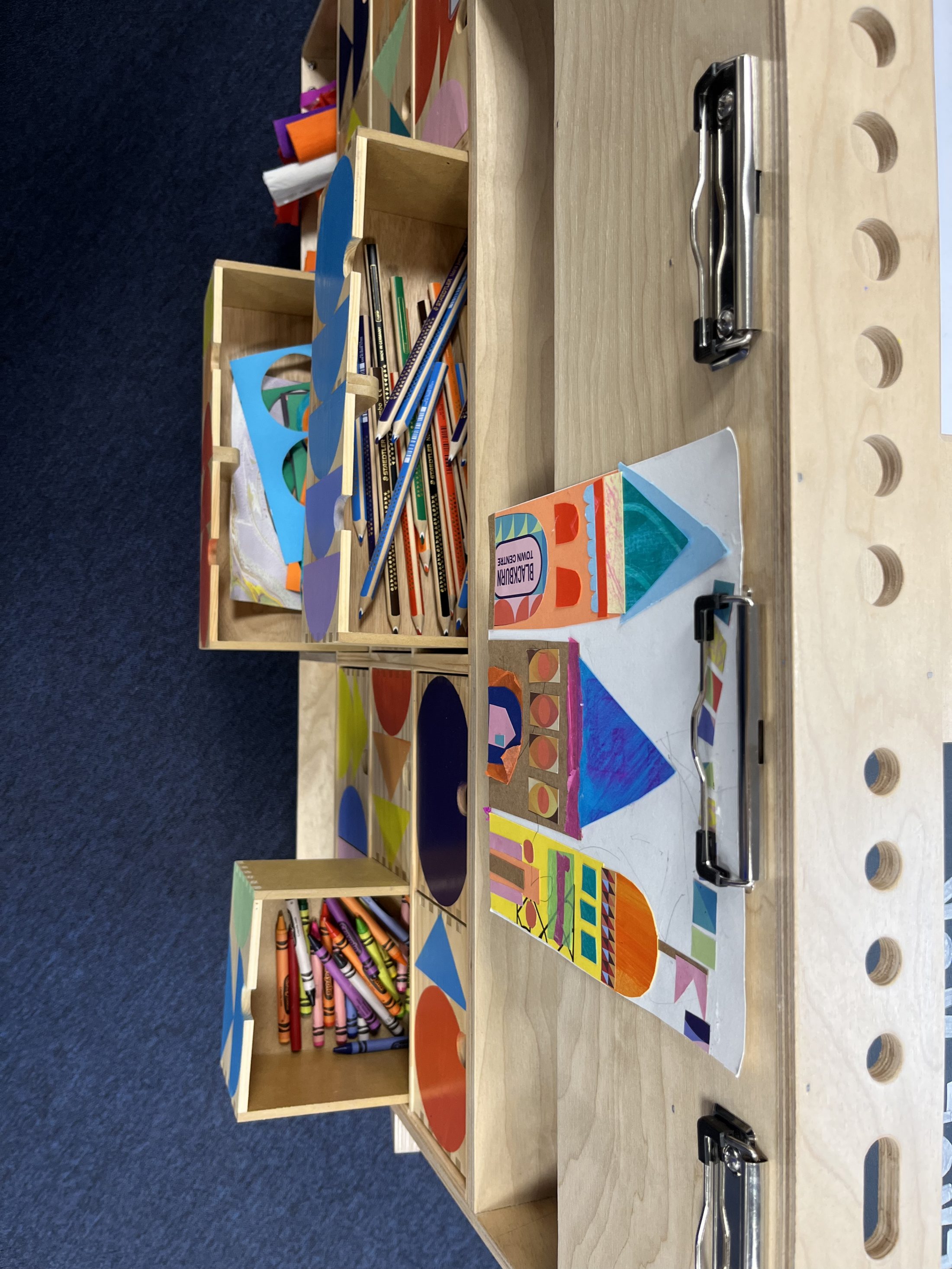 A wooden organiser holds boxes of coloured pencils and crayons. Next to them are colourful geometric art projects on paper, resting on clipboards atop a light wood table, with a dark blue carpet visible beneath.