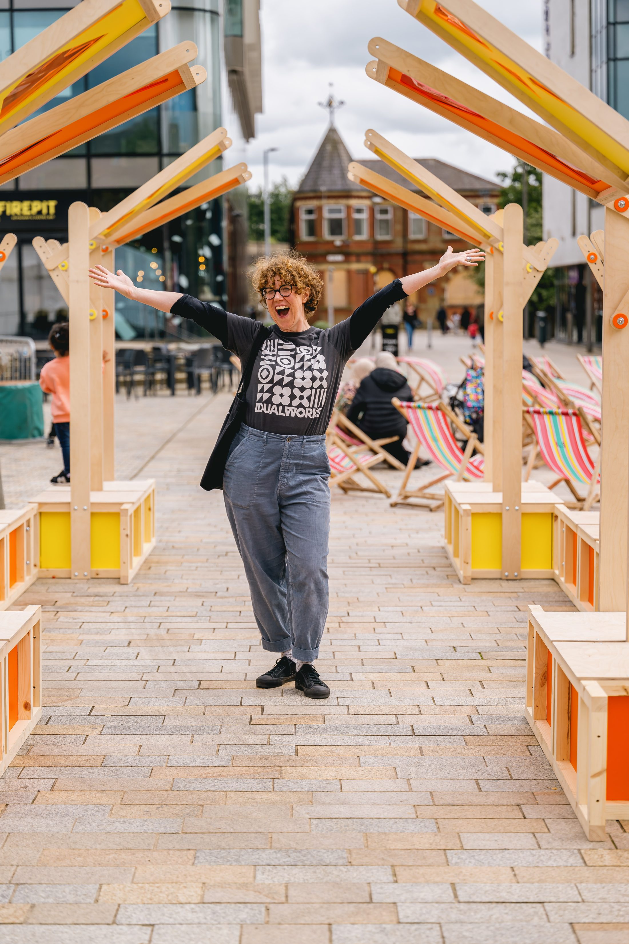 A person with curly hair, glasses, and a black-and-white printed shirt stands smiling with arms raised under a wooden archway on a city street; deckchairs and buildings are visible in the background.