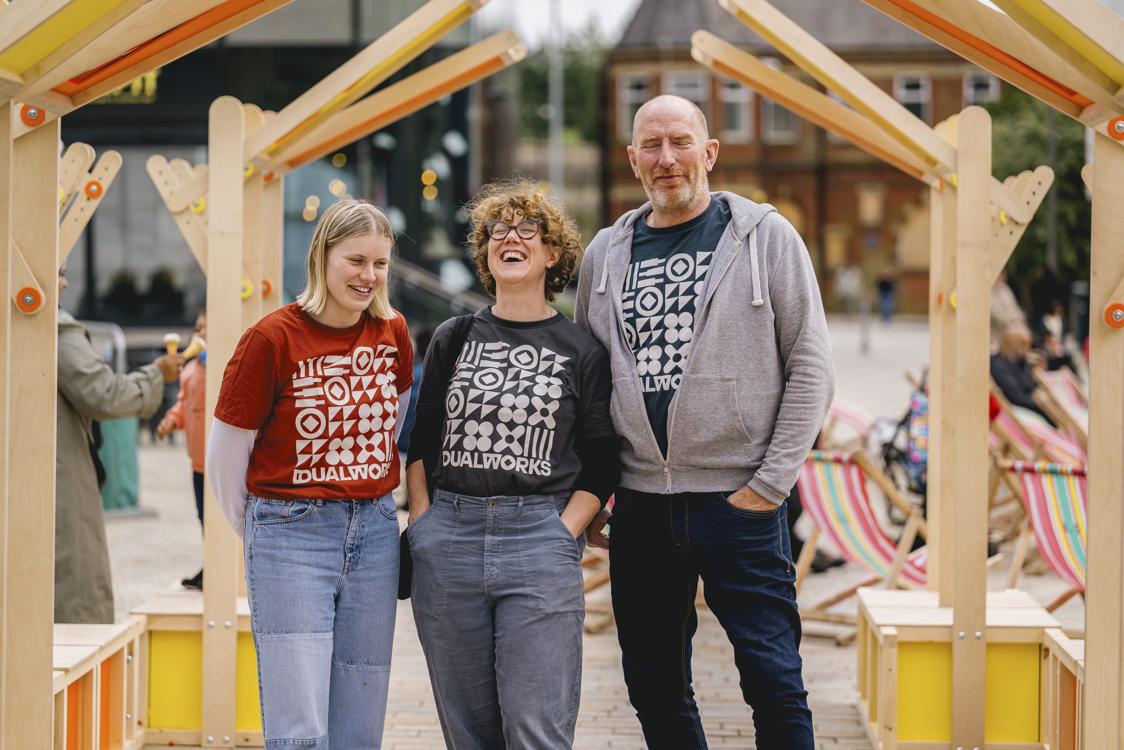 Three adults stand smiling under a wooden arch outdoors. All wear shirts with a bold geometric pattern and the word “DUALWORKS.” Deckchairs and buildings are visible in the background.