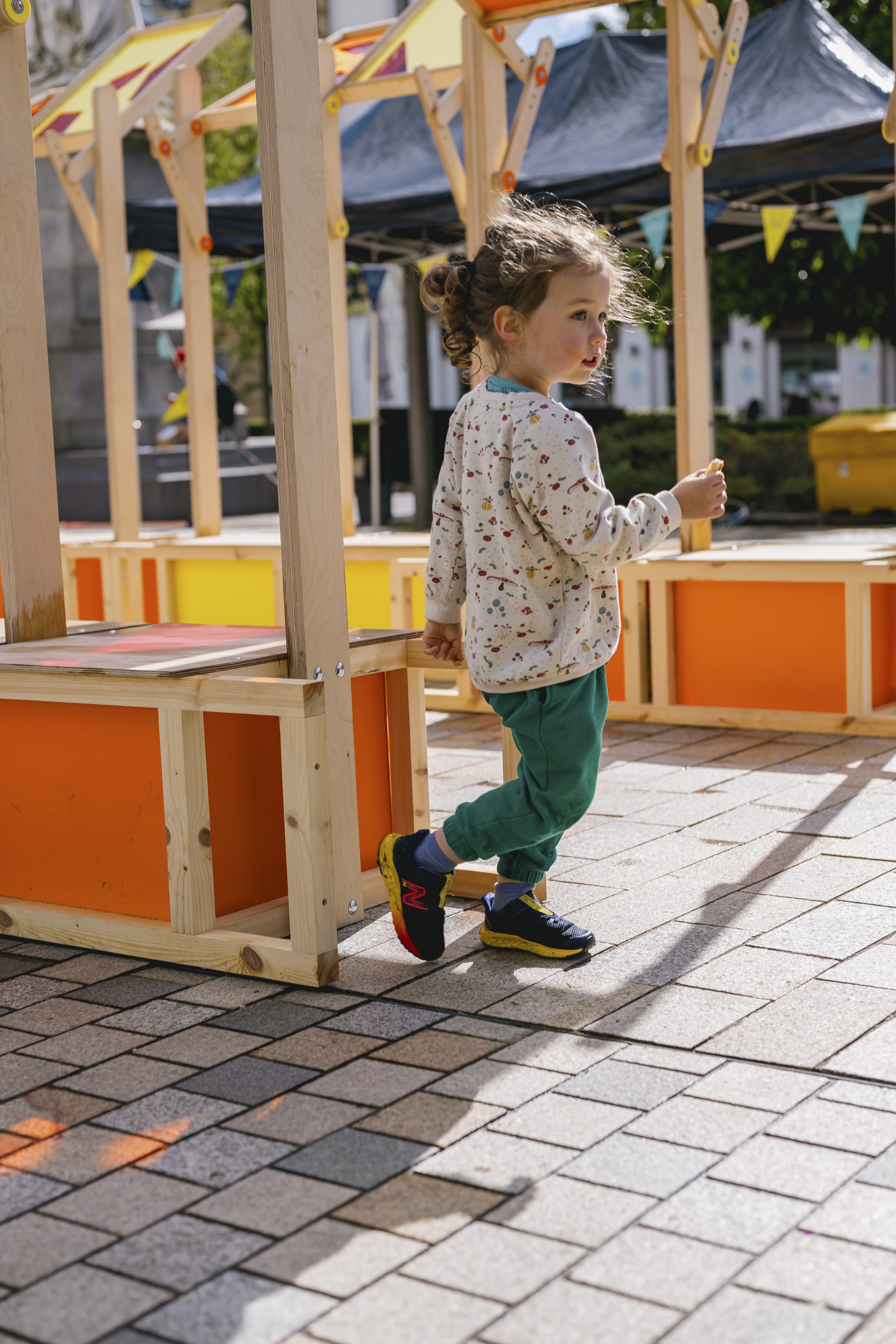 A young child with curly hair, wearing a patterned jumper and green trousers, walks on a paved surface near colourful wooden structures at an outdoor event.