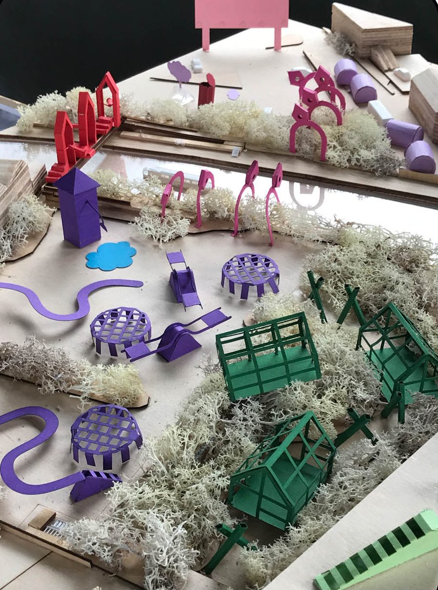A close-up of a colourful architectural model features green, purple, and pink miniature structures, pathways, and grassy textures, resembling a playful park or garden layout with various abstract shapes and forms.