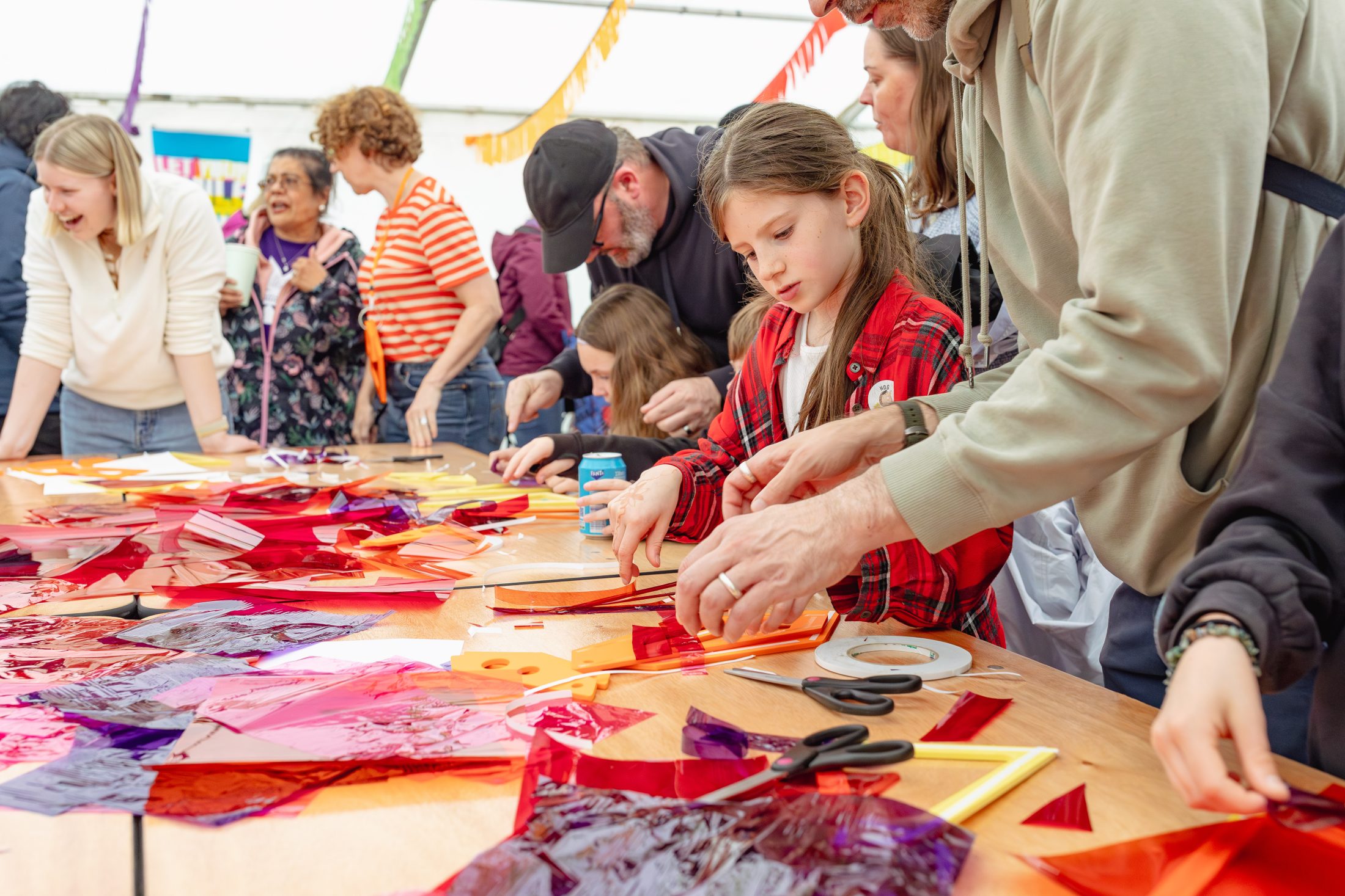 Children and adults gather around a table covered with colourful translucent papers, scissors, and tape, engaging in a hands-on craft activity at a lively community event.