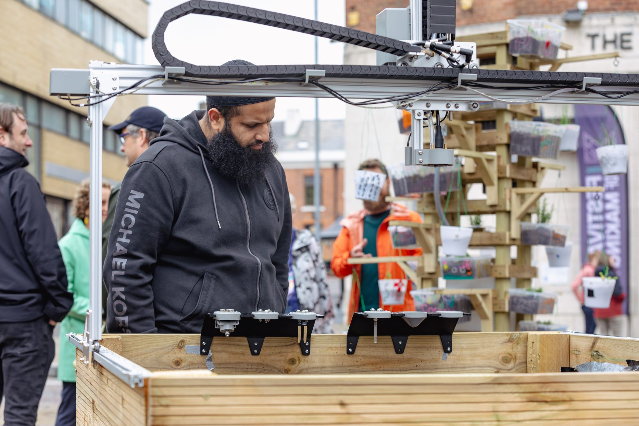 A man wearing a black hoodie stands beside a wooden planter with gardening equipment, observing an urban gardening setup at an outdoor event. People and plant displays are visible in the background.