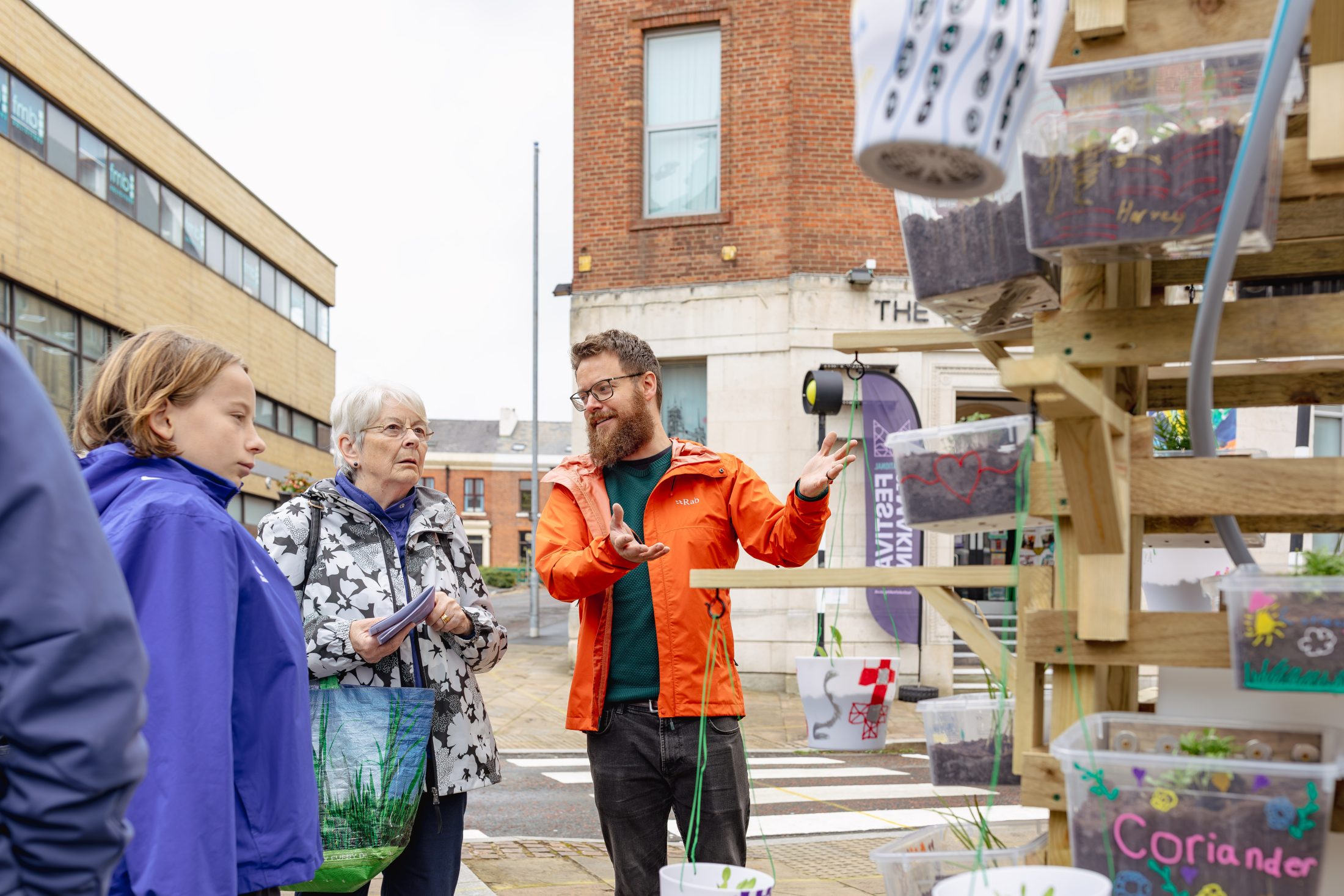 A man in an orange jacket explains a vertical garden display to two women on a city street, with labelled containers of plants like coriander and radish visible.