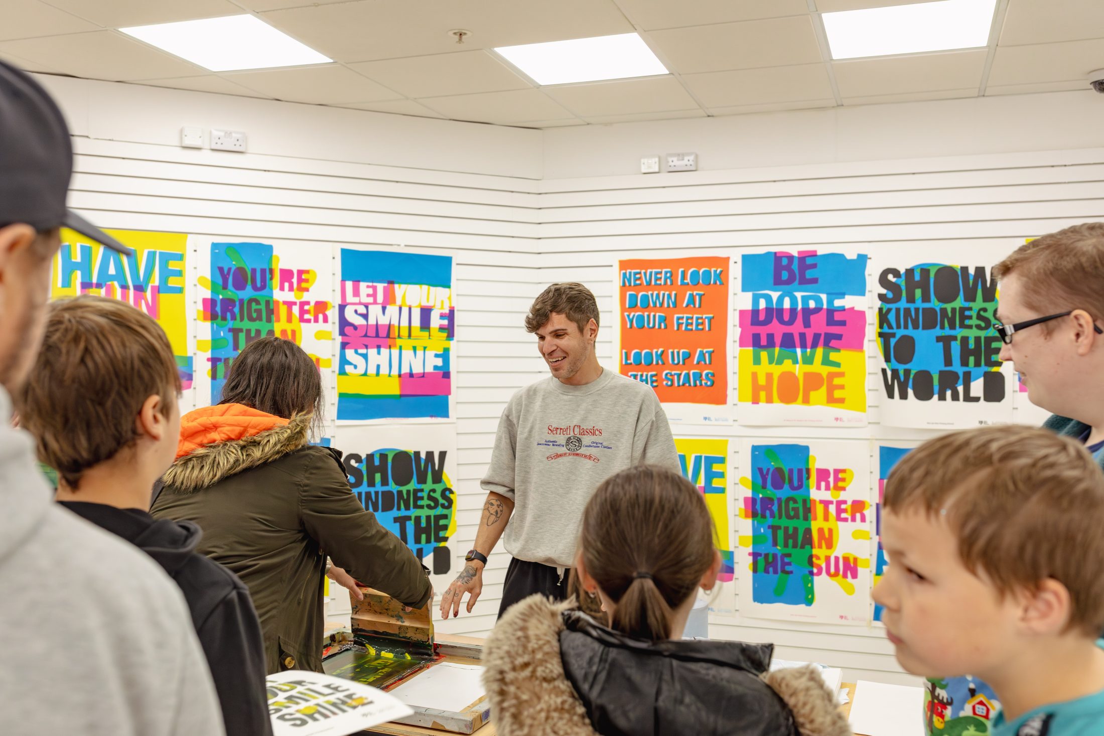 A man demonstrates screen printing to a group of children and adults in a room with colourful, motivational posters on the wall. The posters feature bold messages like “Show Kindness to the World” and “LET YOUR SMILE SHINE.”.