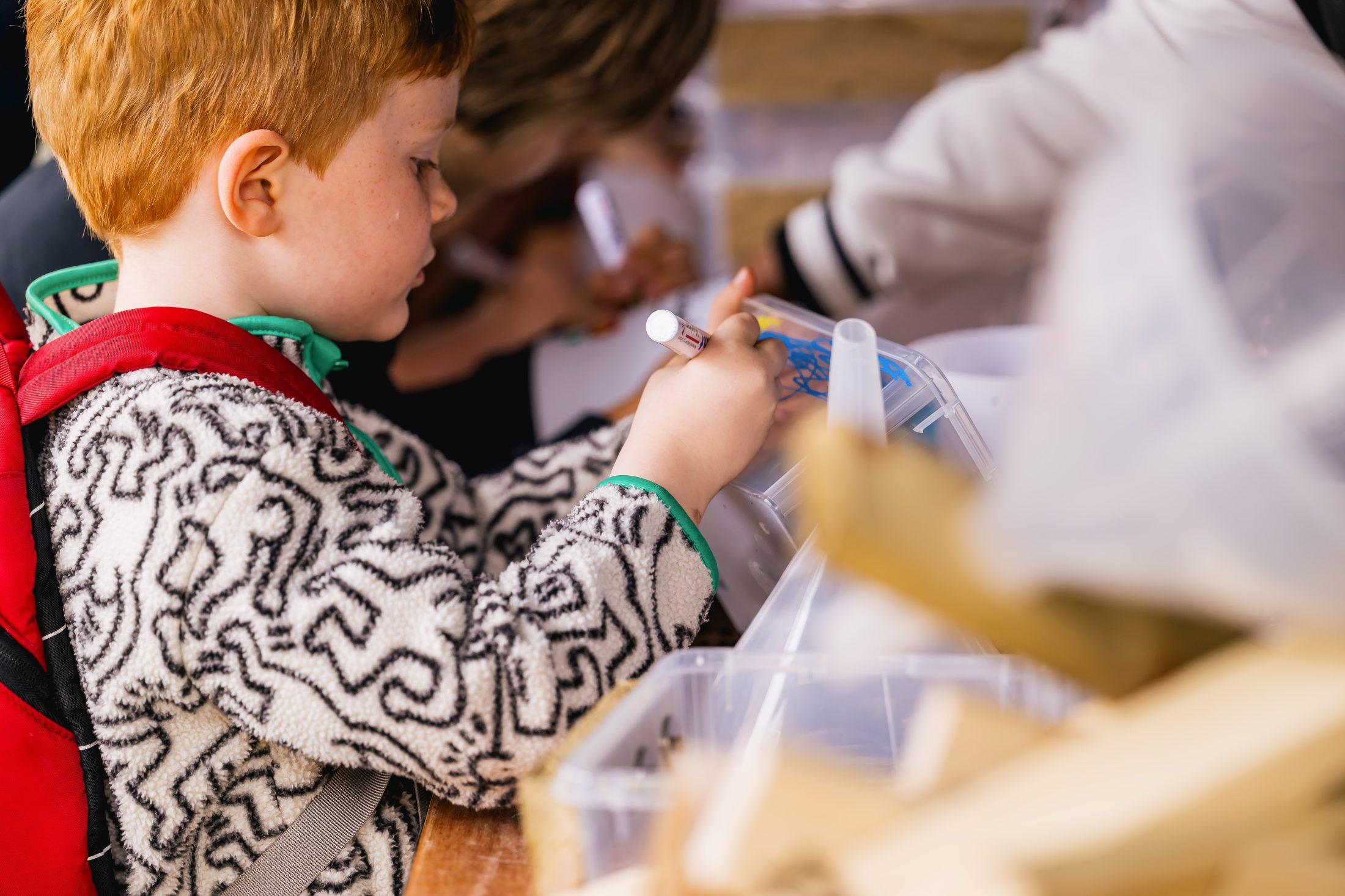 A young child with red hair and a red rucksack draws on a clear plastic object with a marker while sitting at a table with art supplies. Other children are visible in the background out of focus.