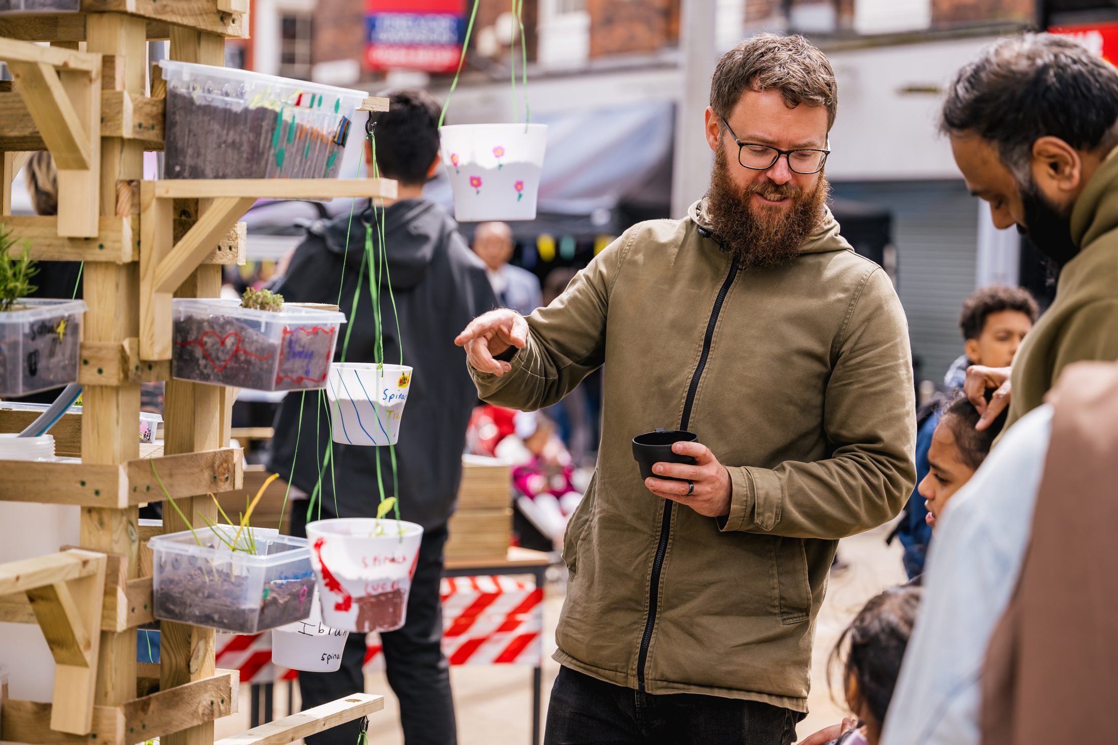 A bearded man with glasses talks to a group of people at an outdoor market stall, surrounded by hanging potted plants in clear containers, with buildings and other shoppers in the background.