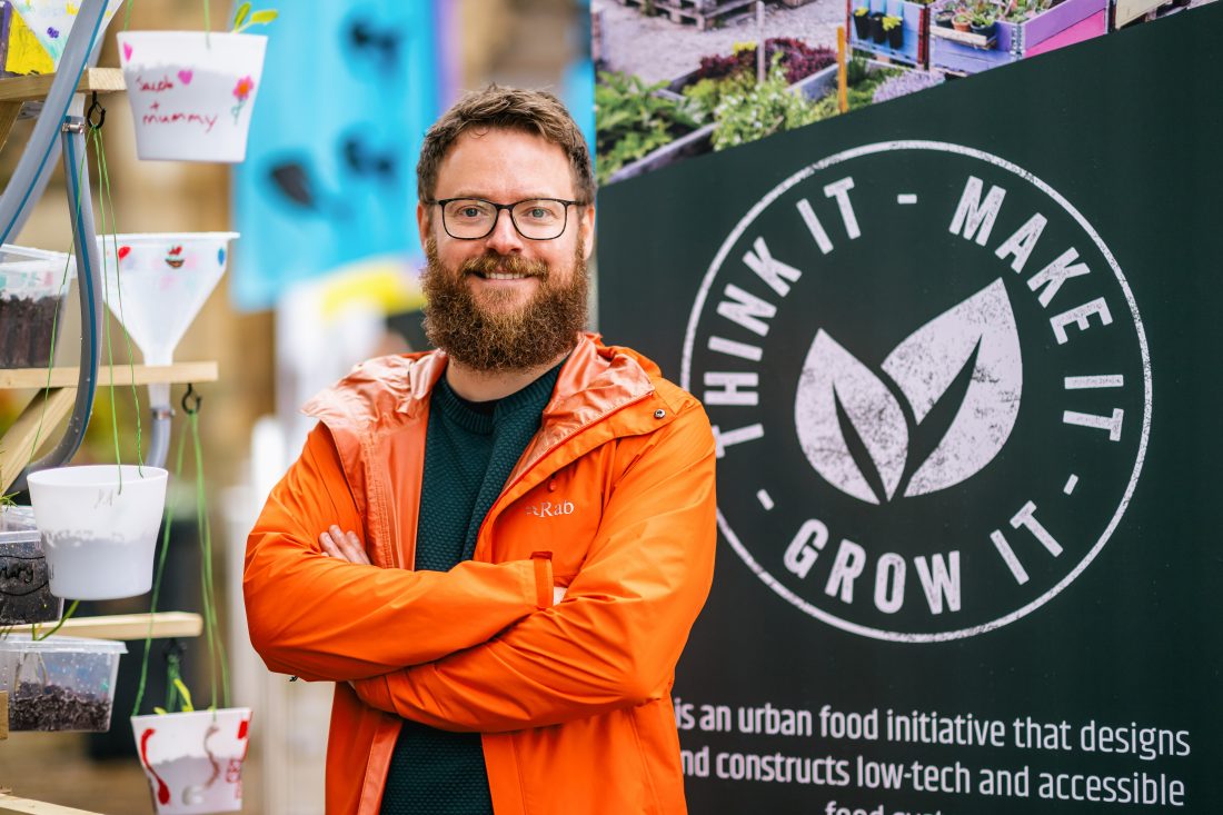 A bearded man in glasses and an orange jacket stands smiling with arms crossed in front of a sign that reads Think It Make It Grow It next to potted plants.