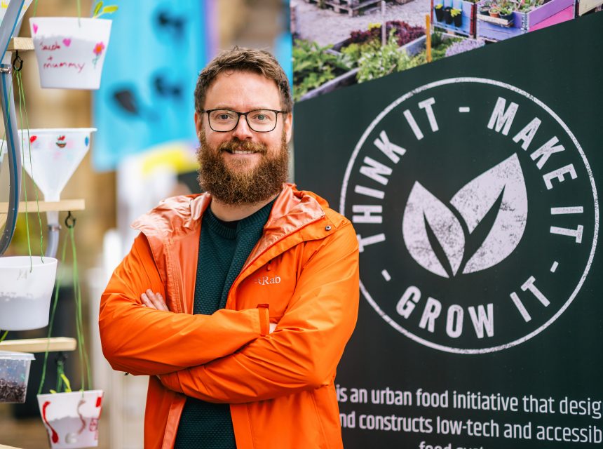 A bearded man in glasses and an orange jacket stands smiling with arms crossed in front of a sign that reads Think It Make It Grow It next to potted plants.