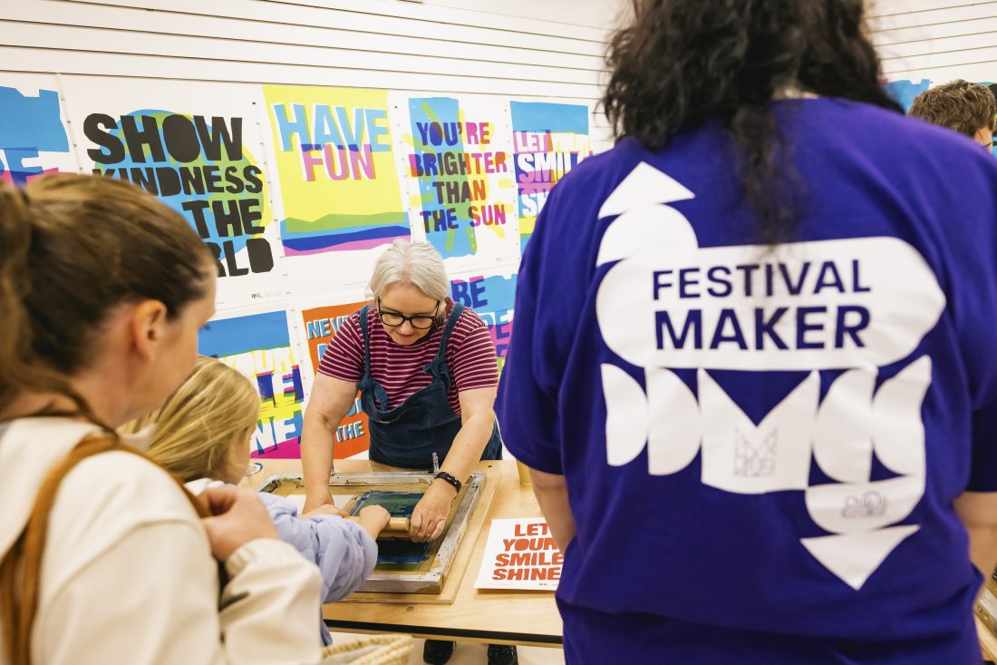 People gather around a table where a person in striped dungarees demonstrates screen printing. Colourful posters with motivational phrases are displayed on the wall. A person in a Festival Maker shirt stands in the foreground.