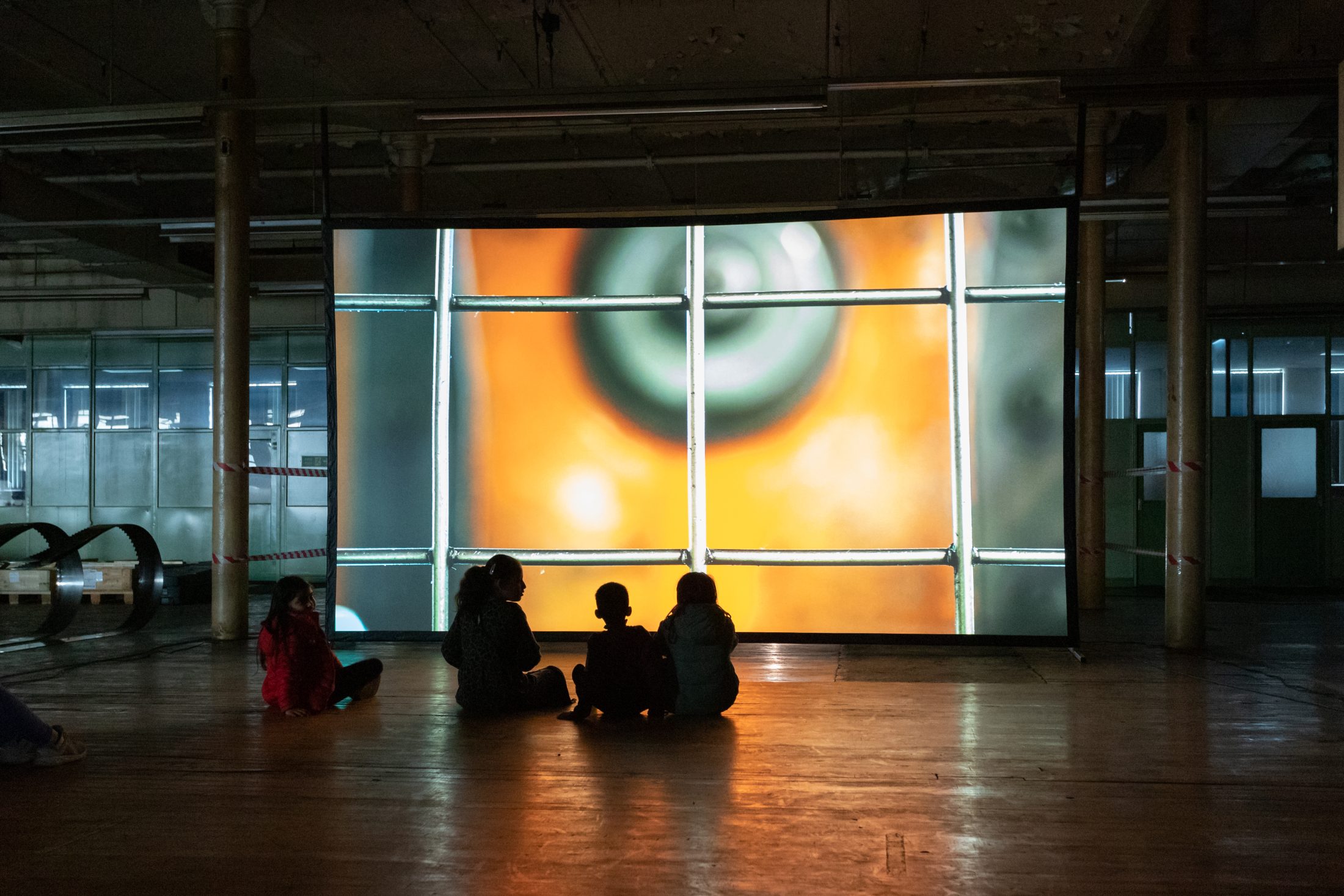 Four children sit on the floor in a dimly lit room, watching a large screen displaying a close-up image with metal bars and a circular object with orange and green tones.