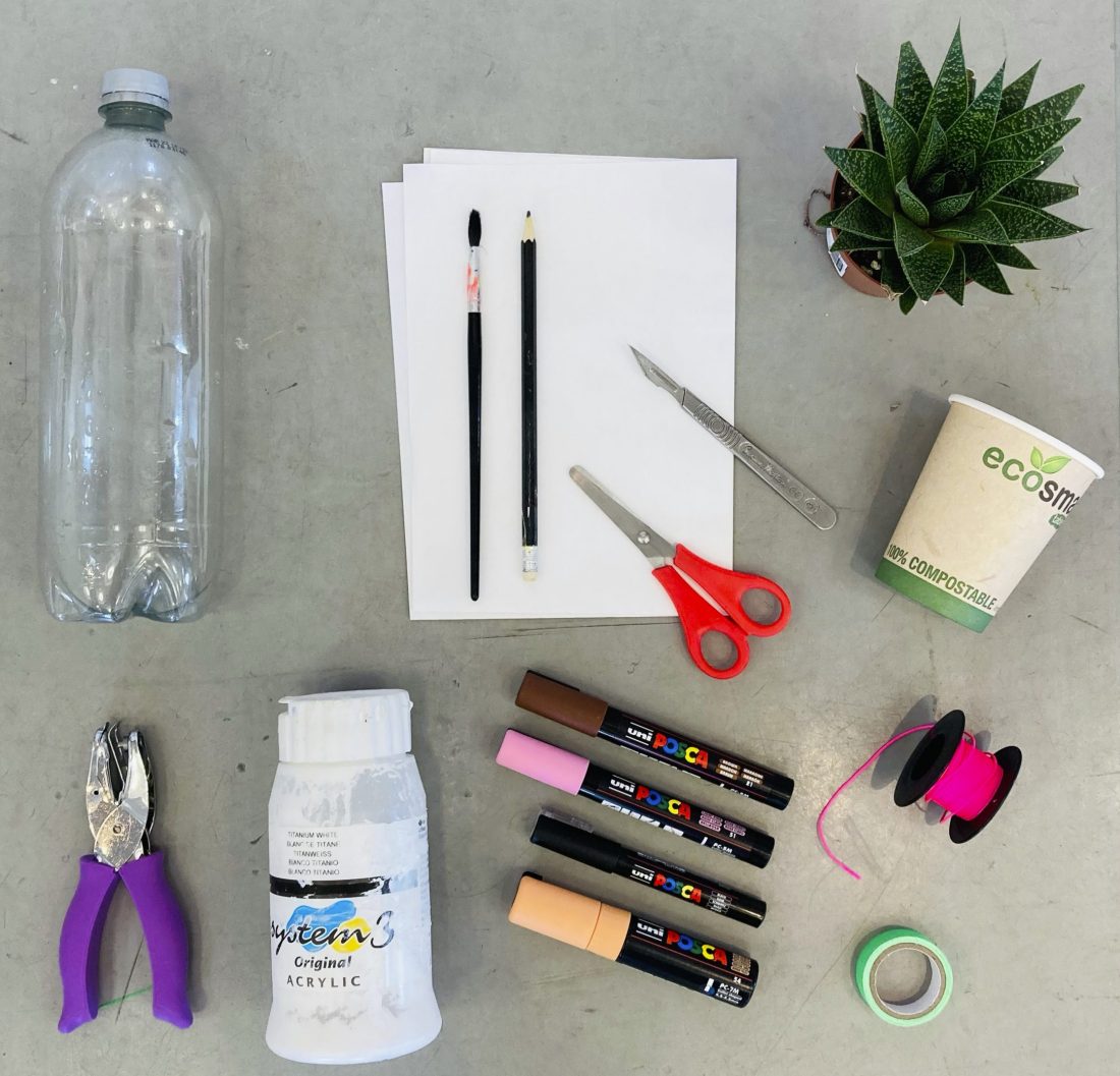 A flat lay of art and craft supplies: empty plastic bottle, hole punch, acrylic gesso, two paintbrushes, paper sheets, scissors, tweezers, four paint markers, tape, string, compostable cup, and a small potted plant.
